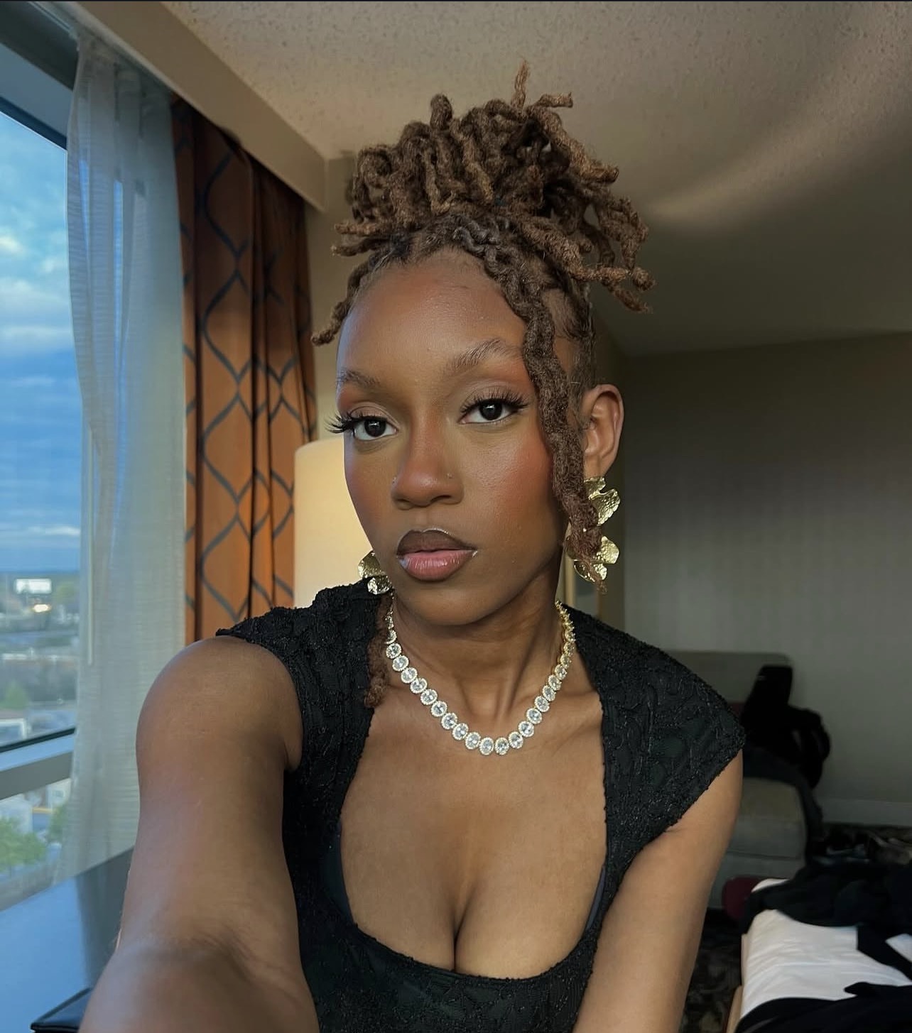 Young woman with styled hair, wearing a black top, pearl necklace, and earrings, in a room with window and curtains.
