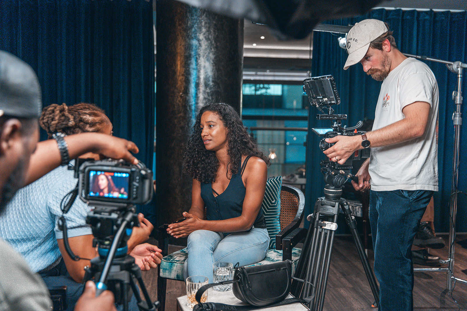 A woman sits in front of a camera crew during an interview, with a cameraman filming her, in an indoor setting.