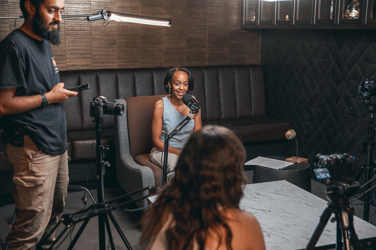 Woman sitting in a chair speaking into a microphone during a recording session, with two people operating cameras and equipment.
