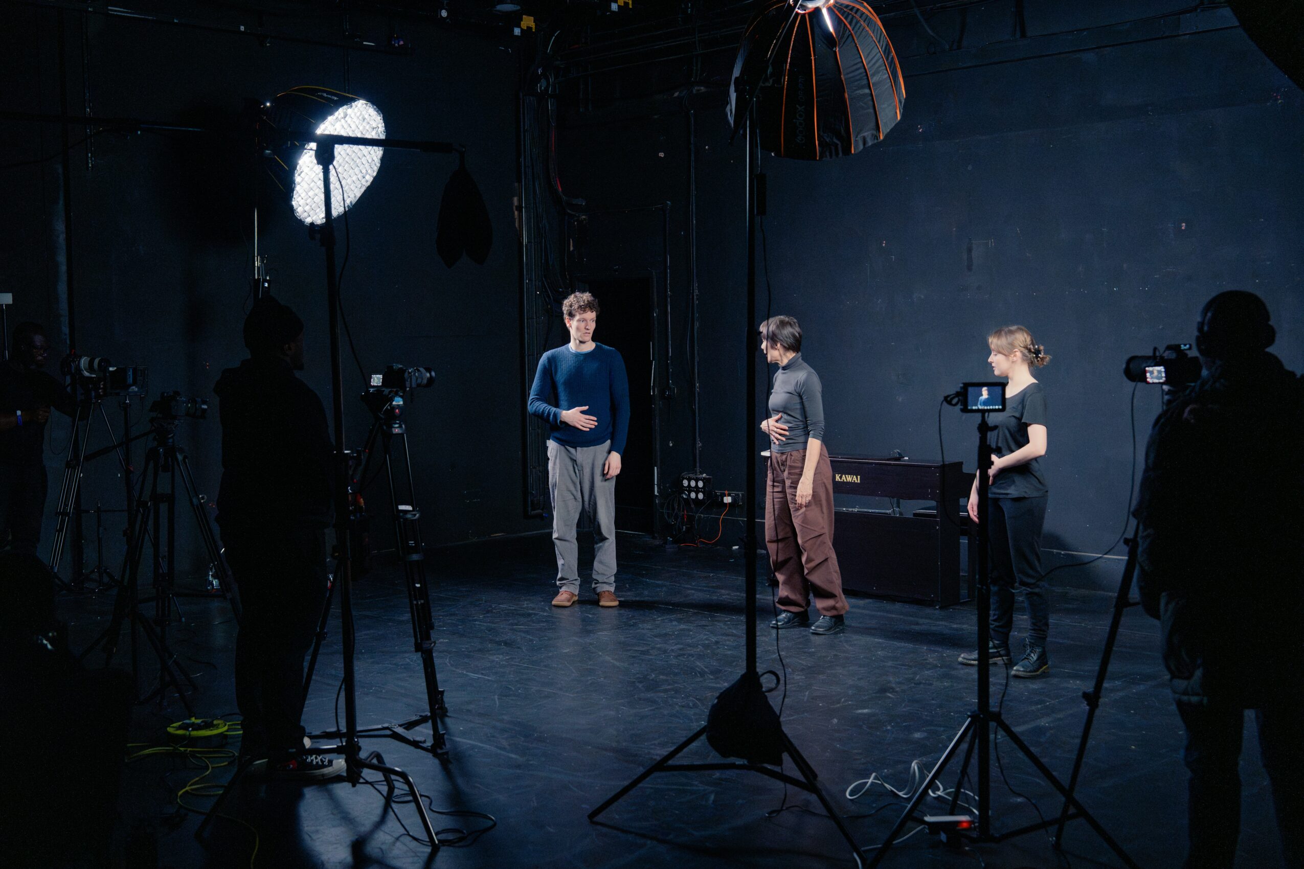 Three people stand in a dark studio with professional lighting and cameras, preparing for a video or photo shoot.