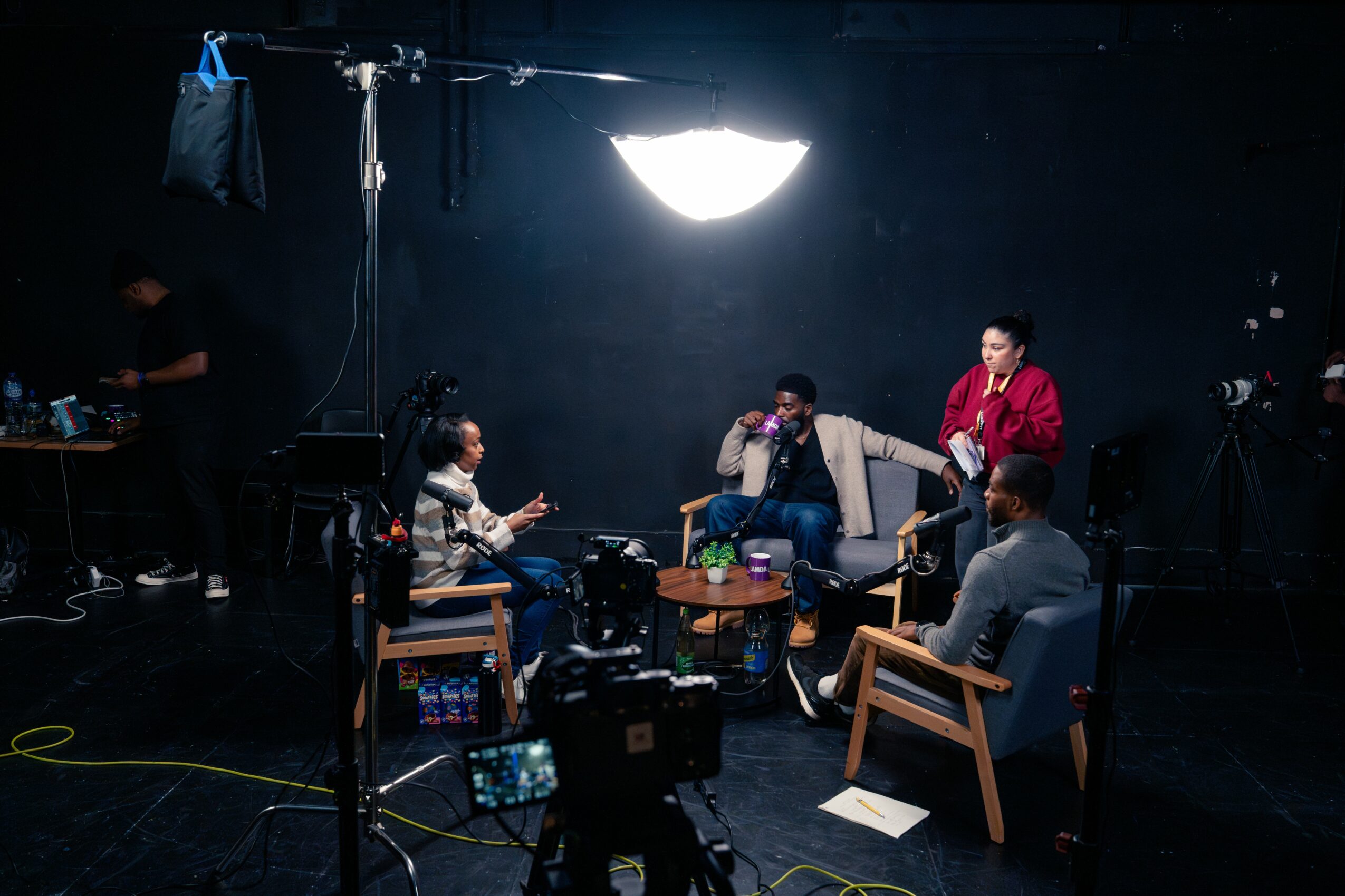 Four people sit around a table in a studio with a large light overhead, filming or discussing, with equipment visible.