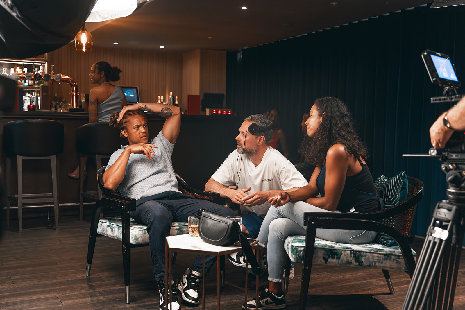 Three people sit and talk in a cafe or lounge, with a bar and a person working behind it in the background.