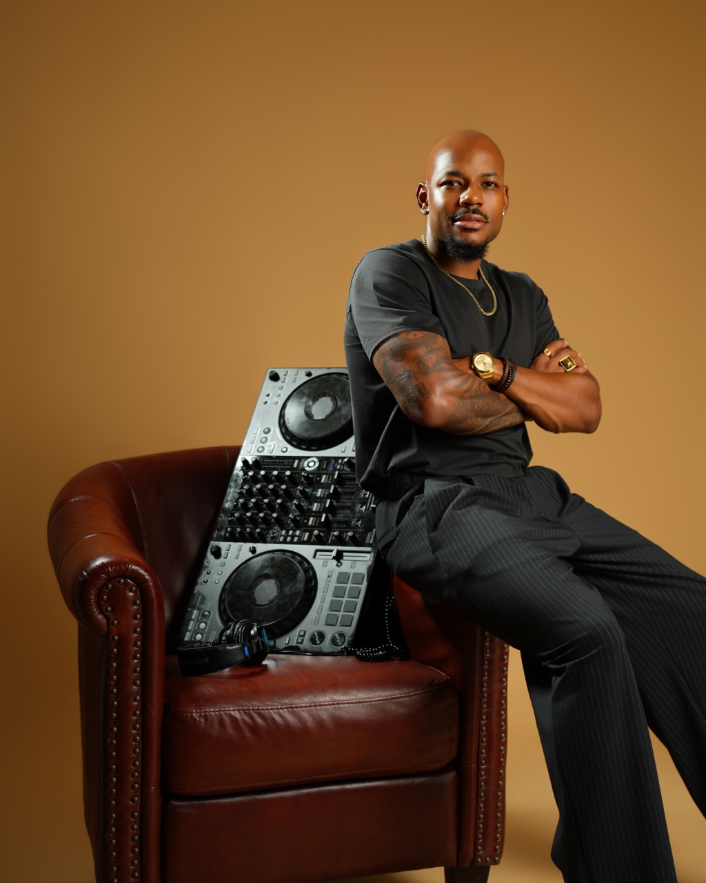 Man with crossed arms sitting on a leather chair, with DJ equipment behind him, against a plain brown background.