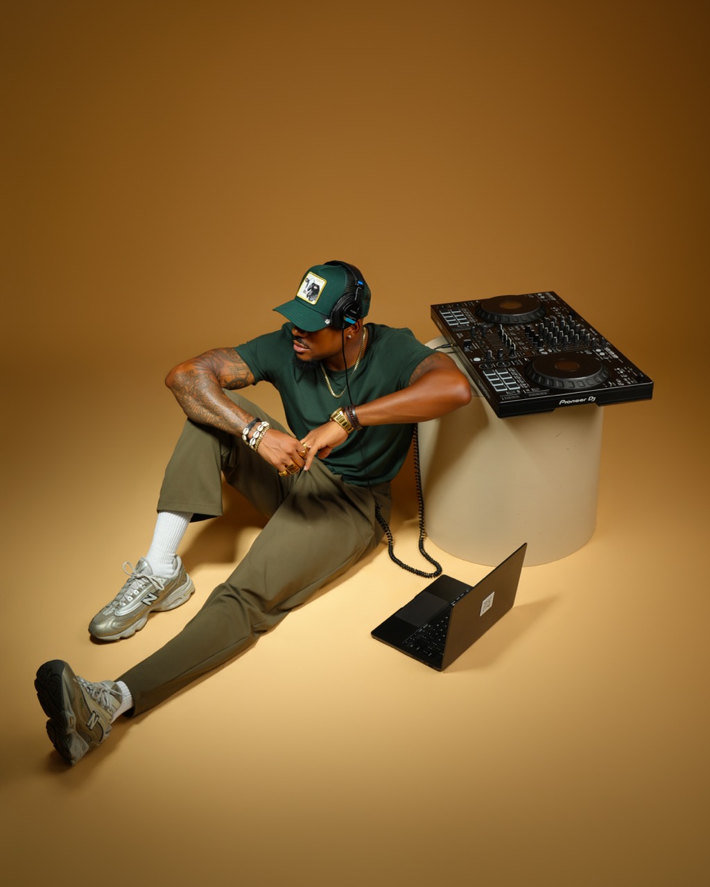 Person sitting on the floor with DJ equipment, wearing a cap, sunglasses, and casual clothing, against a brown background.