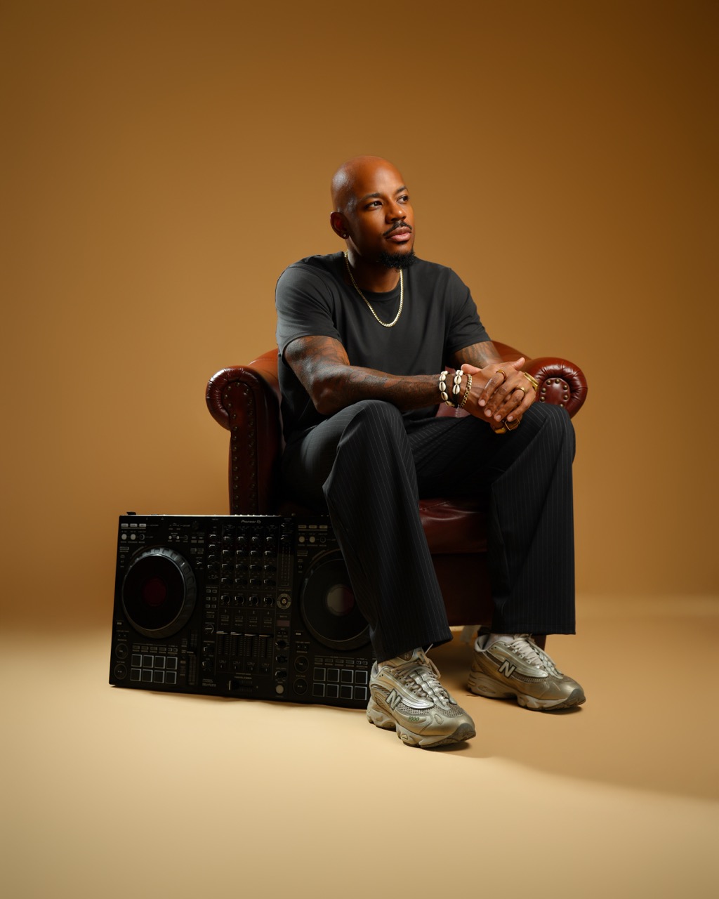 Man sitting on a vintage armchair with a brown background, wearing black clothes and sneakers, with a boombox at his feet.