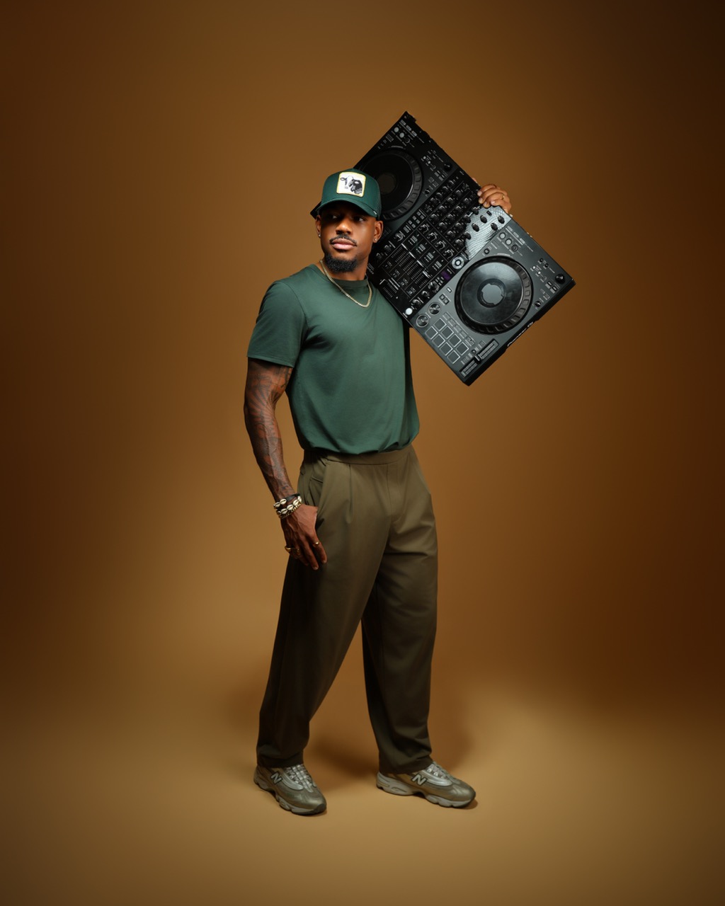 Man wearing a green shirt, brown pants, and a cap, holding DJ equipment on his shoulder, standing against a brown background.