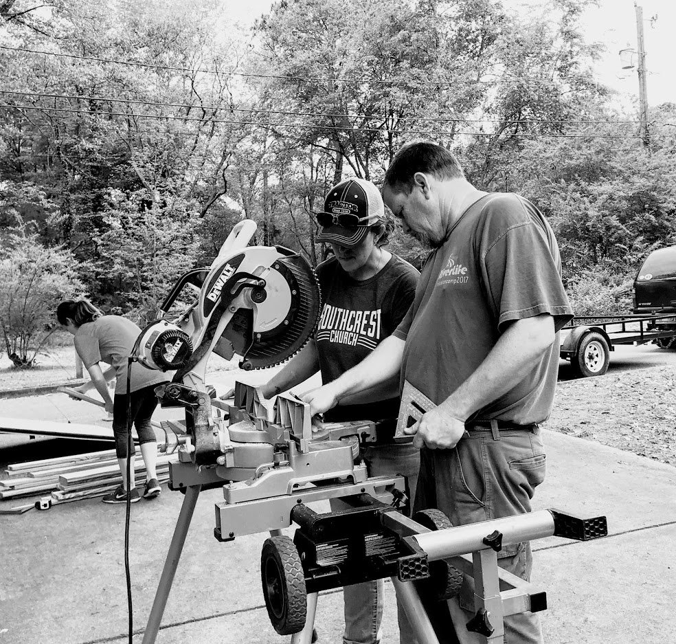 Two men work with a large saw outdoors, with trees and a car in the background.