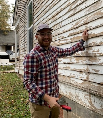 Man smiling, wearing glasses and a cap, standing outside next to a weathered wooden wall, holding a tool.