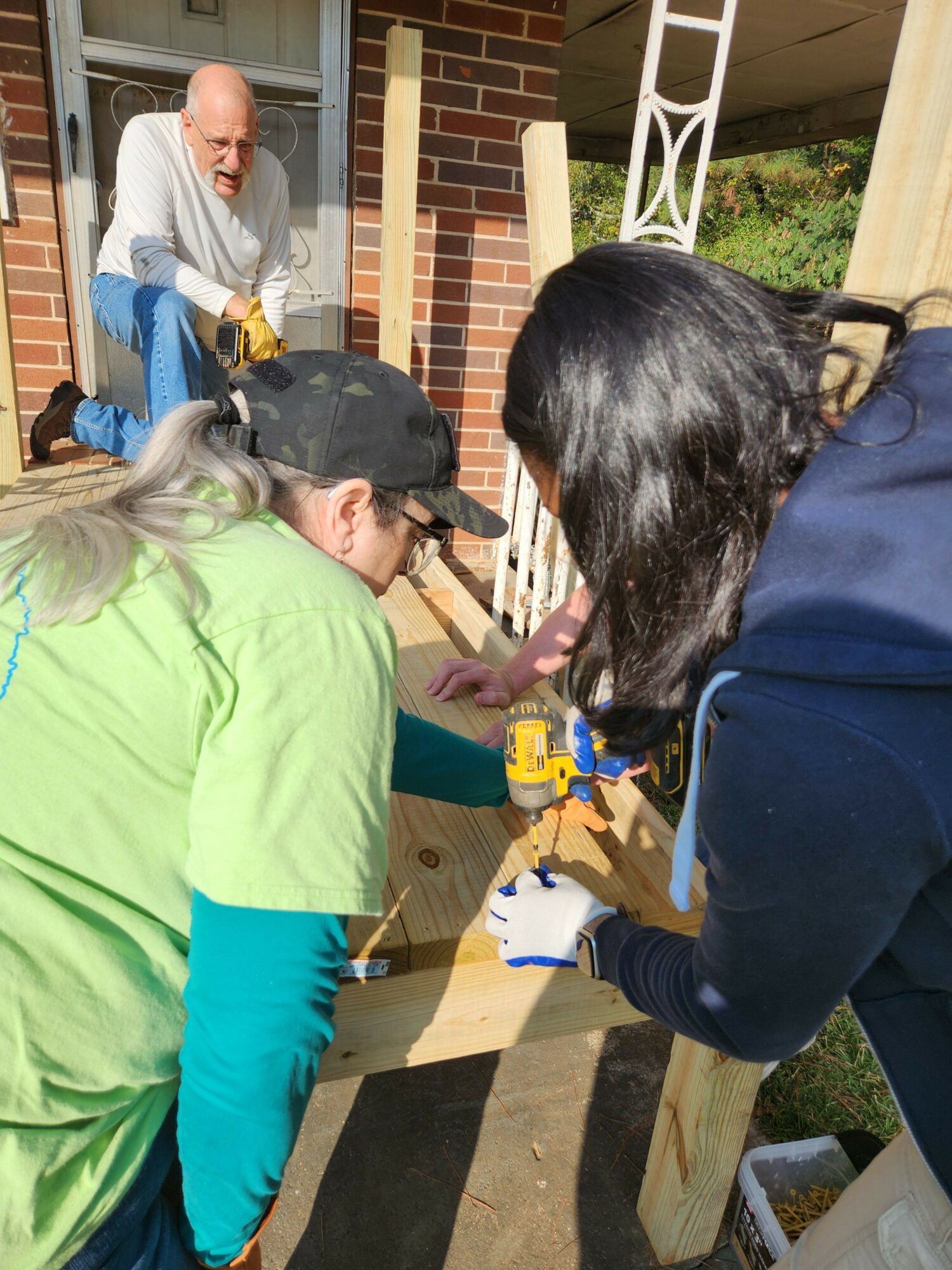 Two women working on a wooden deck, one in a green shirt and the other in a dark jacket, with a person in the background.