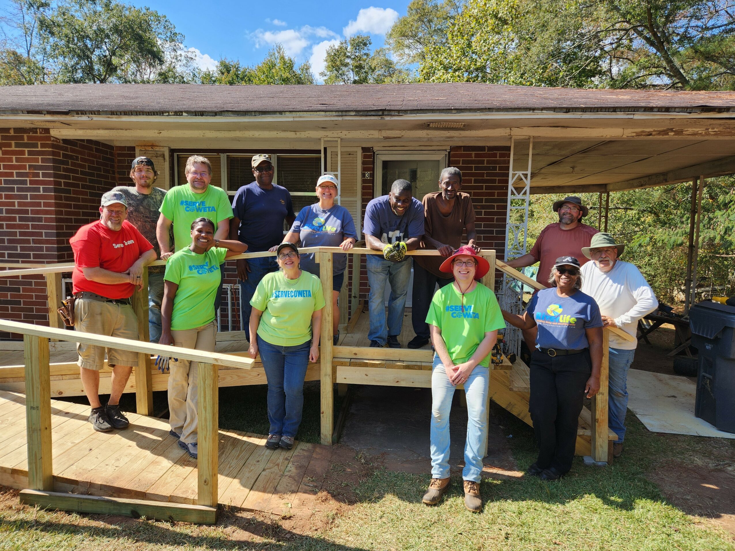 Group of people standing on a wooden porch in front of a brick building, outdoors with trees and blue sky, some wearing bright shirts.