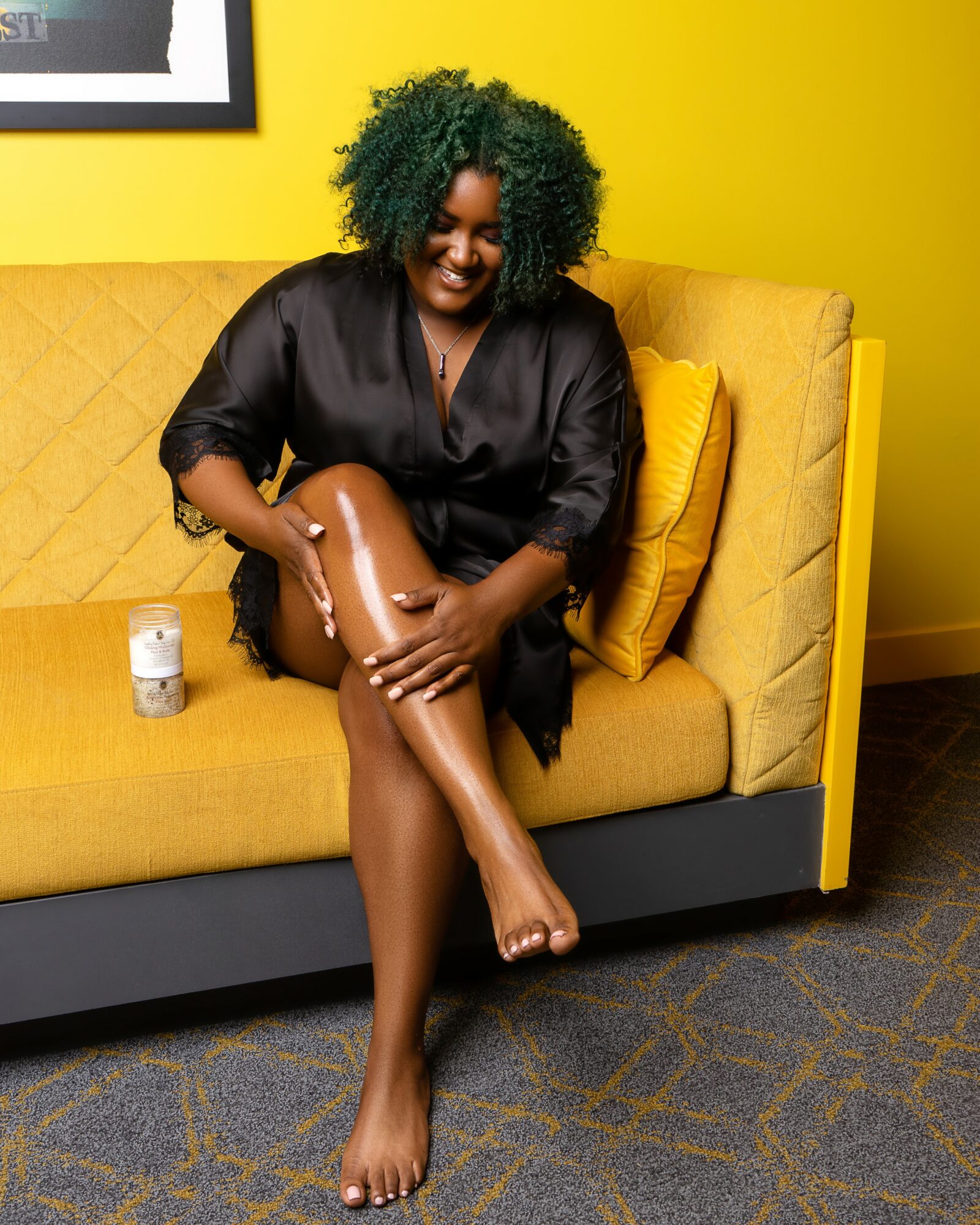 Smiling woman with green curly hair sitting on yellow sofa, holding her leg, with yellow wall behind her.