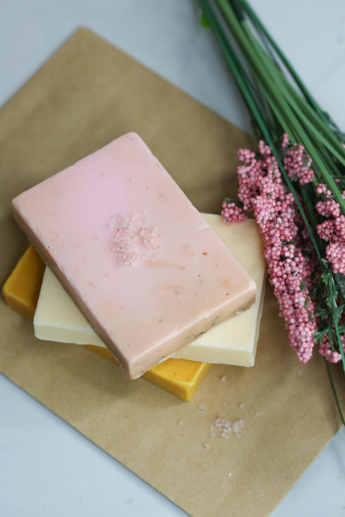 Stack of three soap bars with pink, white, and yellow colors, on brown paper with pink flowers nearby.