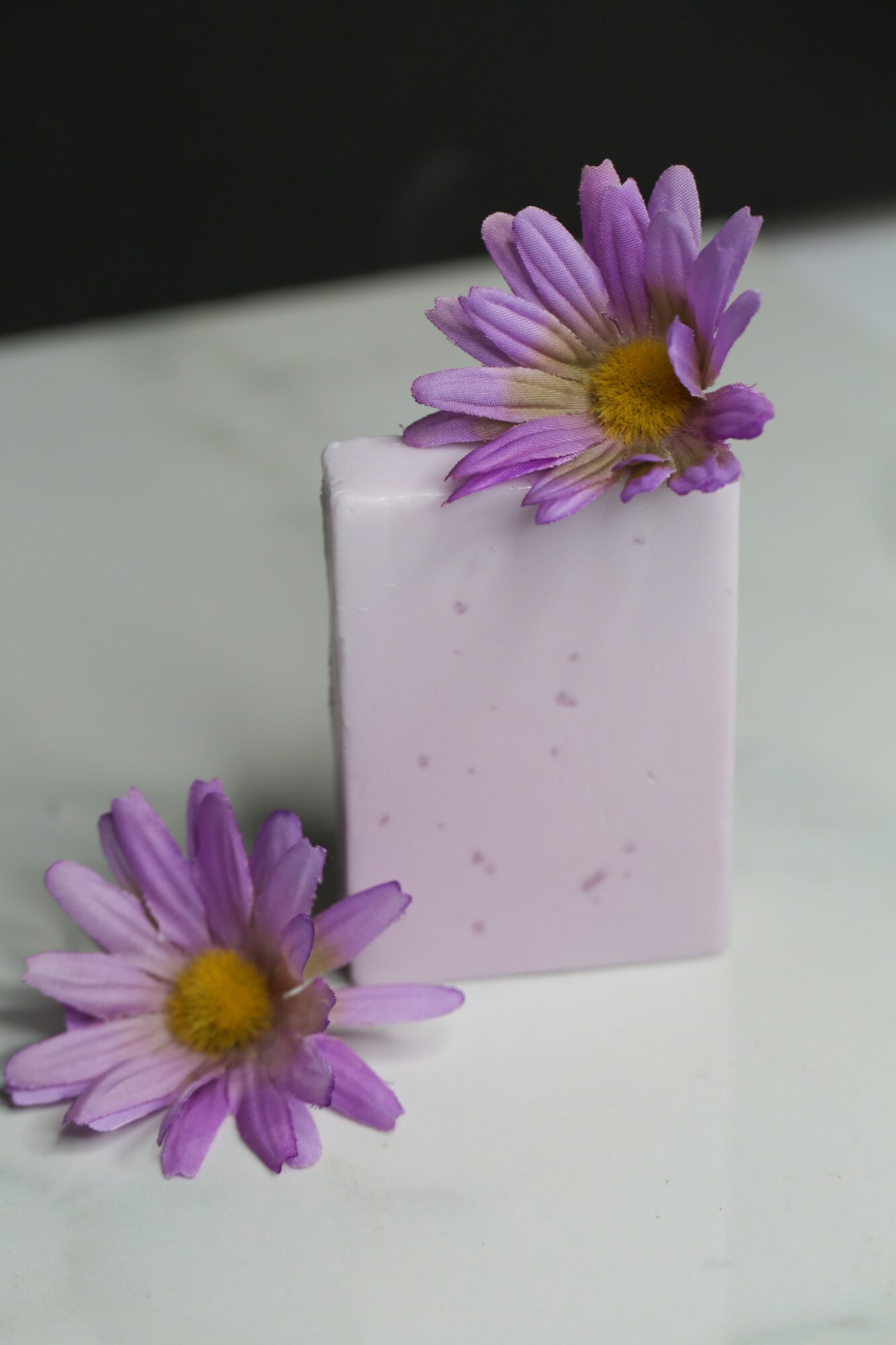 A purple flower with a yellow center rests on a light purple soap bar, with another flower beside it on a white surface.