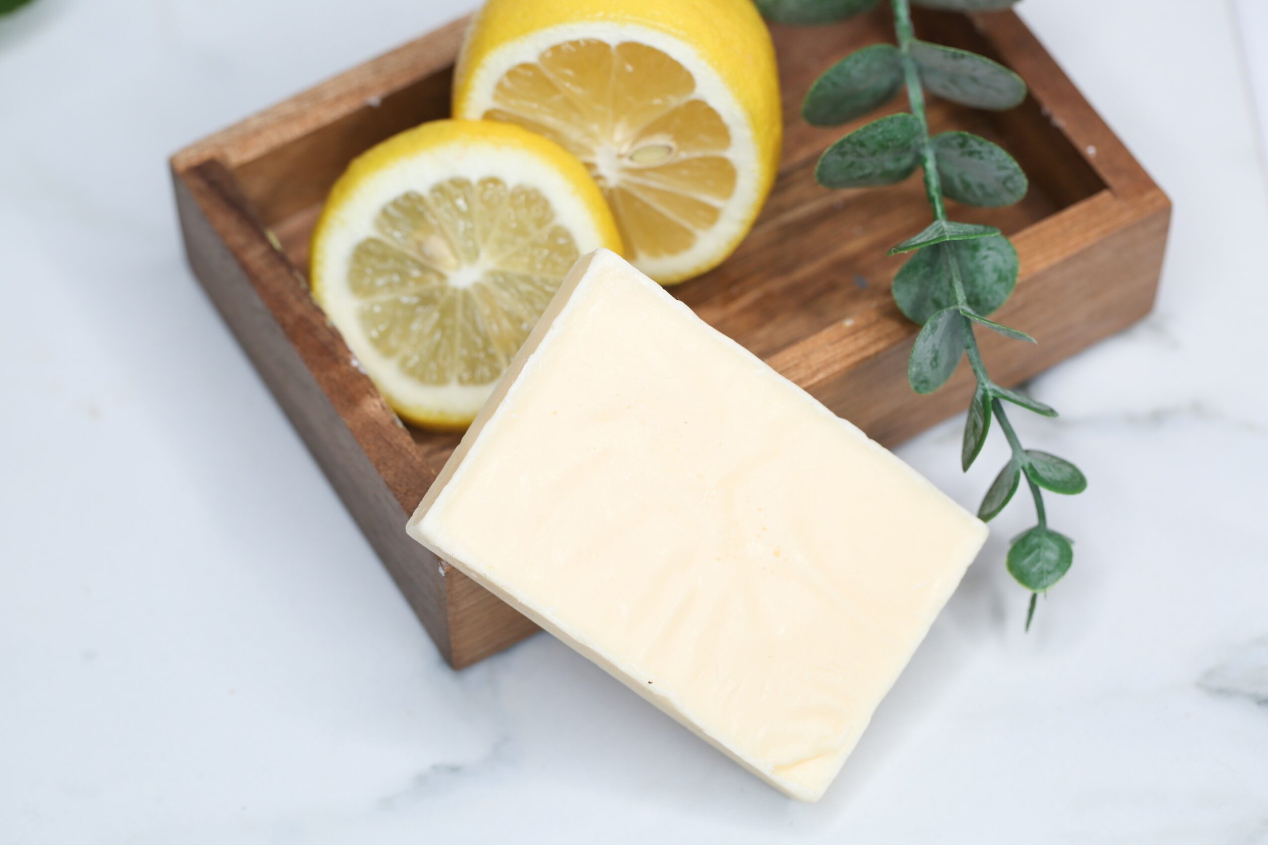 Lemon slices, a bar of soap, and a green plant on a wooden tray, on a white marble surface.