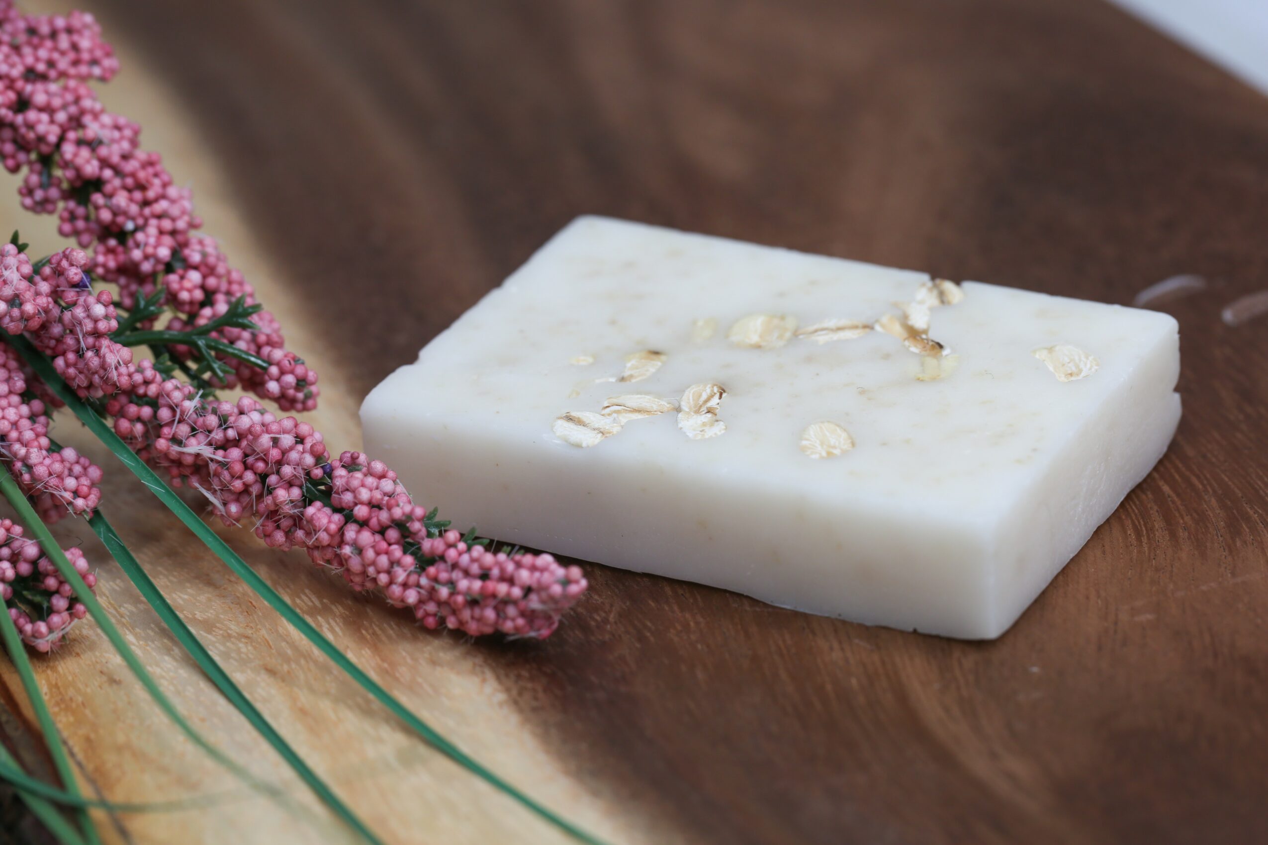 White soap bar with oats on a wooden surface, pink flowers nearby, natural setting.