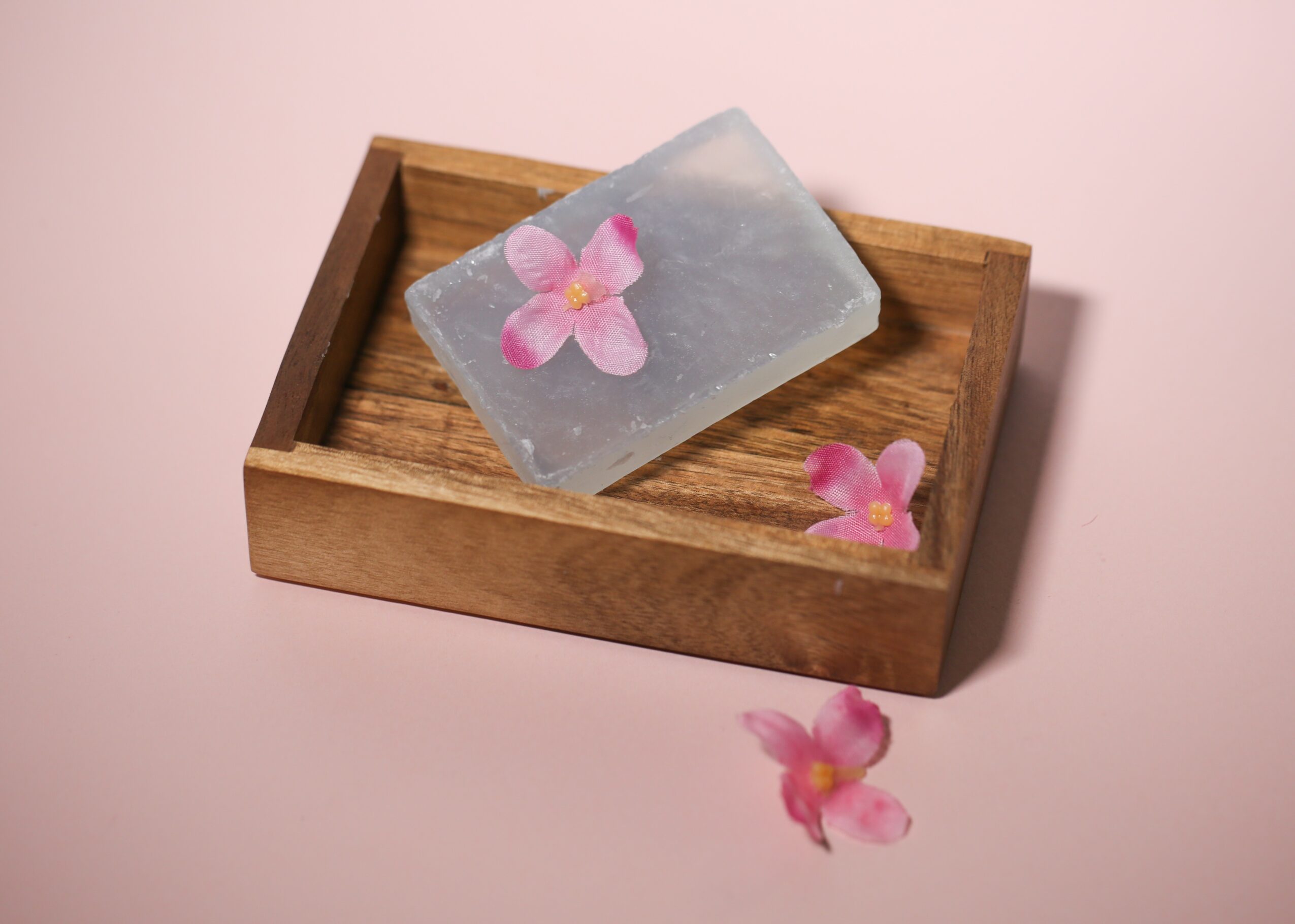 Soap with pink flowers on a wooden tray, with one flower outside the tray, on a pink background.