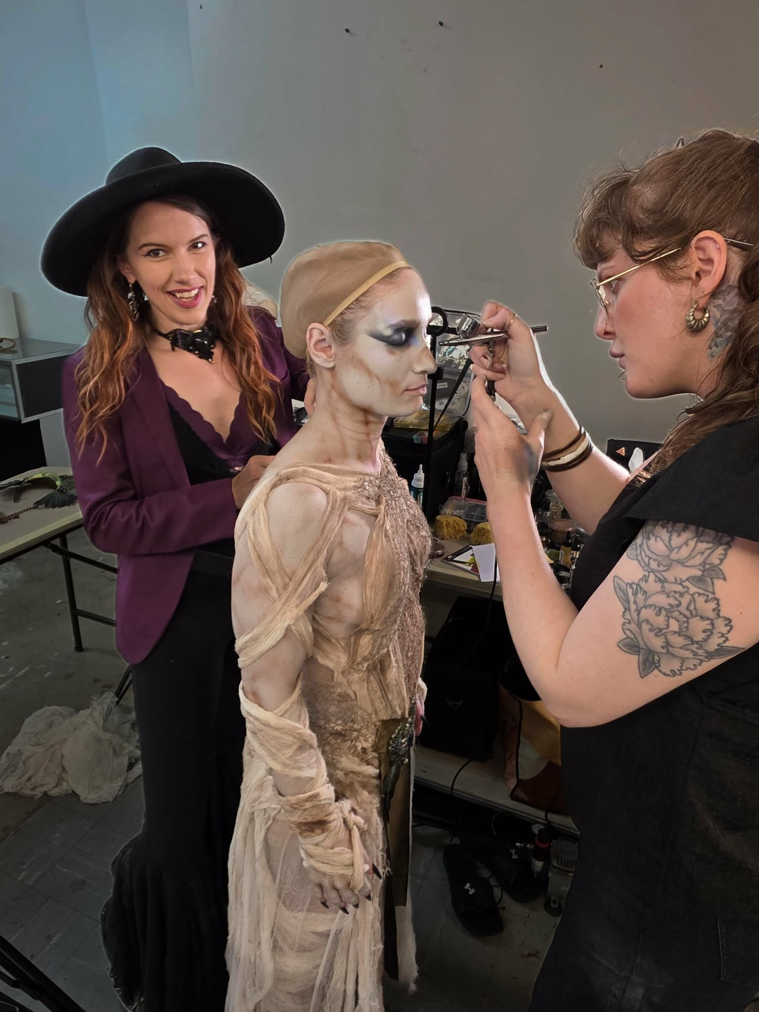 A makeup artist applies makeup to a person in costume, with another person smiling nearby, in a backstage setting.