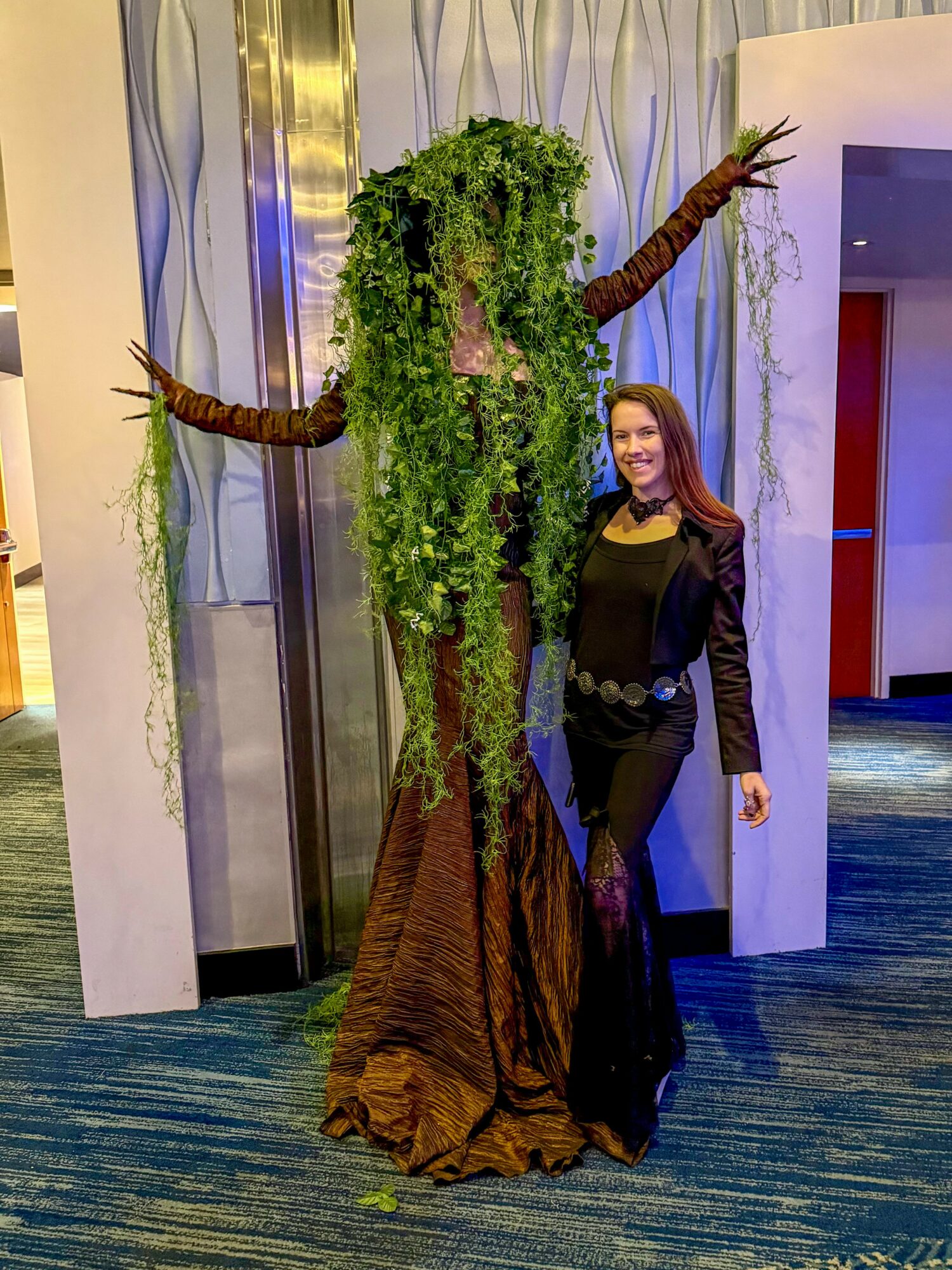 Person standing next to a large tree-like sculpture with green foliage and brown branches, indoors with curtains and a blue carpet.