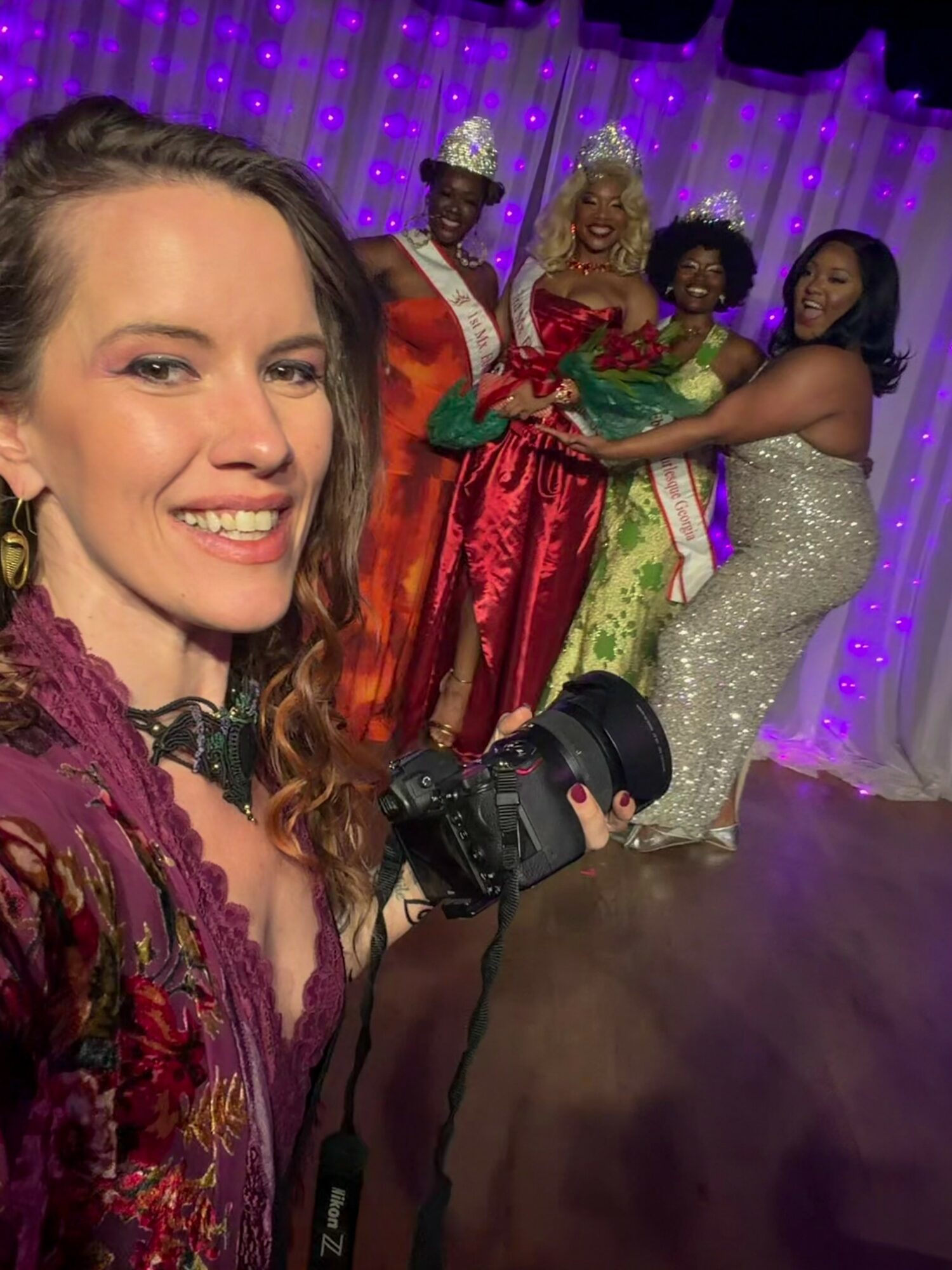 A woman takes a selfie with four women in colorful dresses and crowns, holding a bouquet, in front of purple lights.