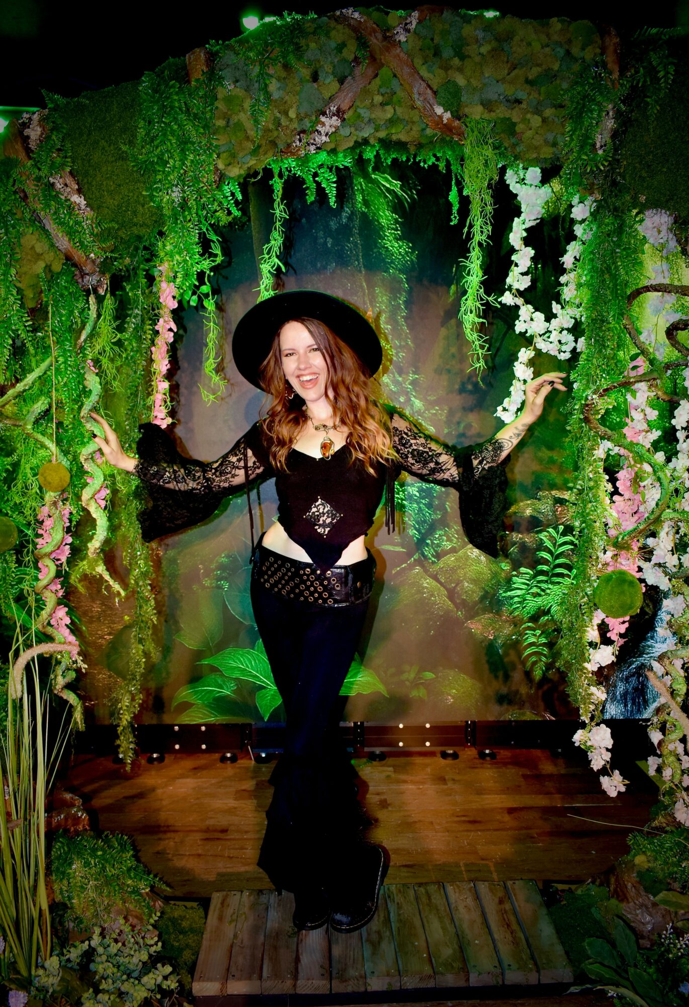 Woman in black outfit and wide-brimmed hat standing in a lush, green, forest-like setting with plants and flowers.