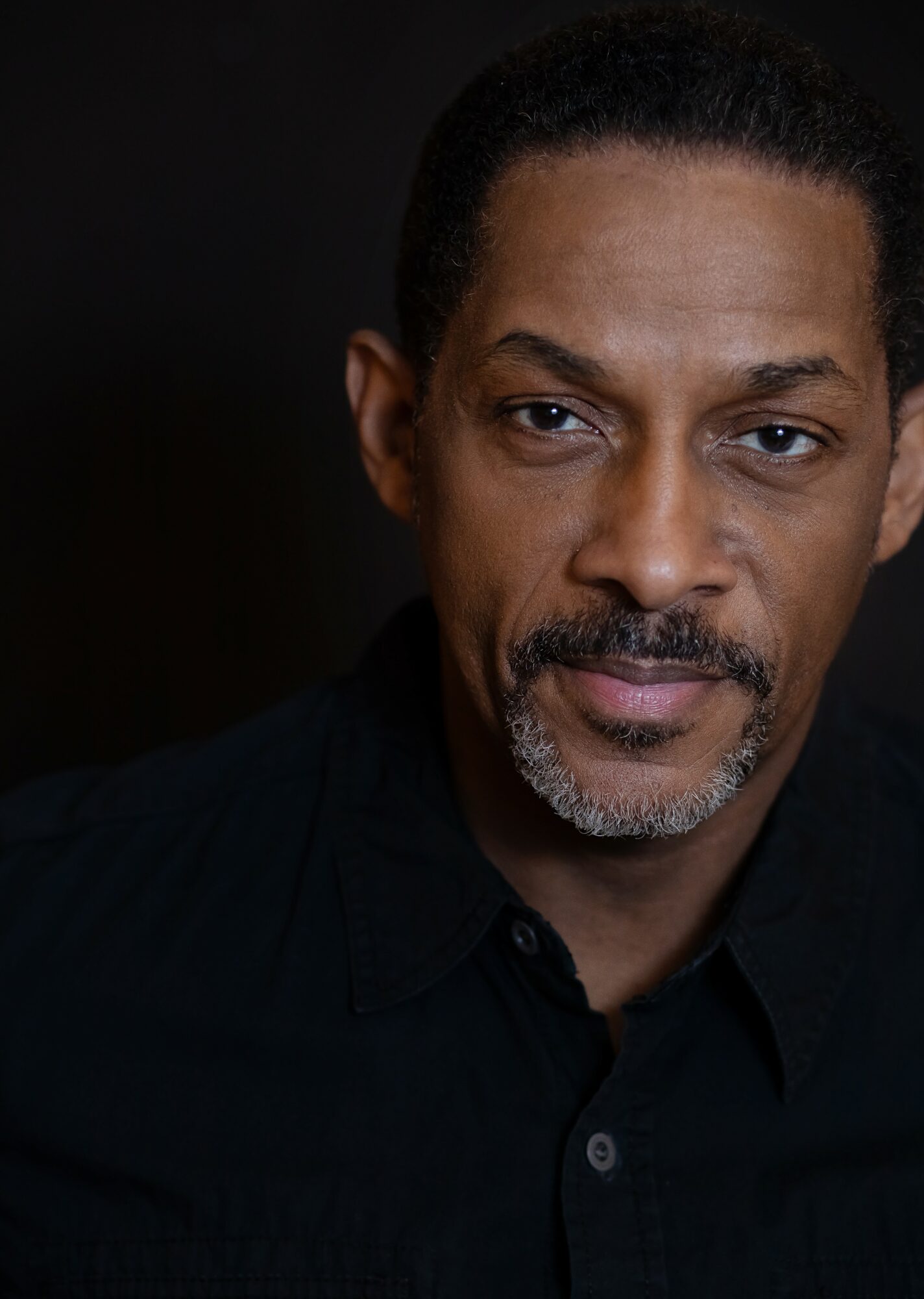 Portrait of a man with short dark hair, a goatee, wearing a black shirt, looking at the camera.
