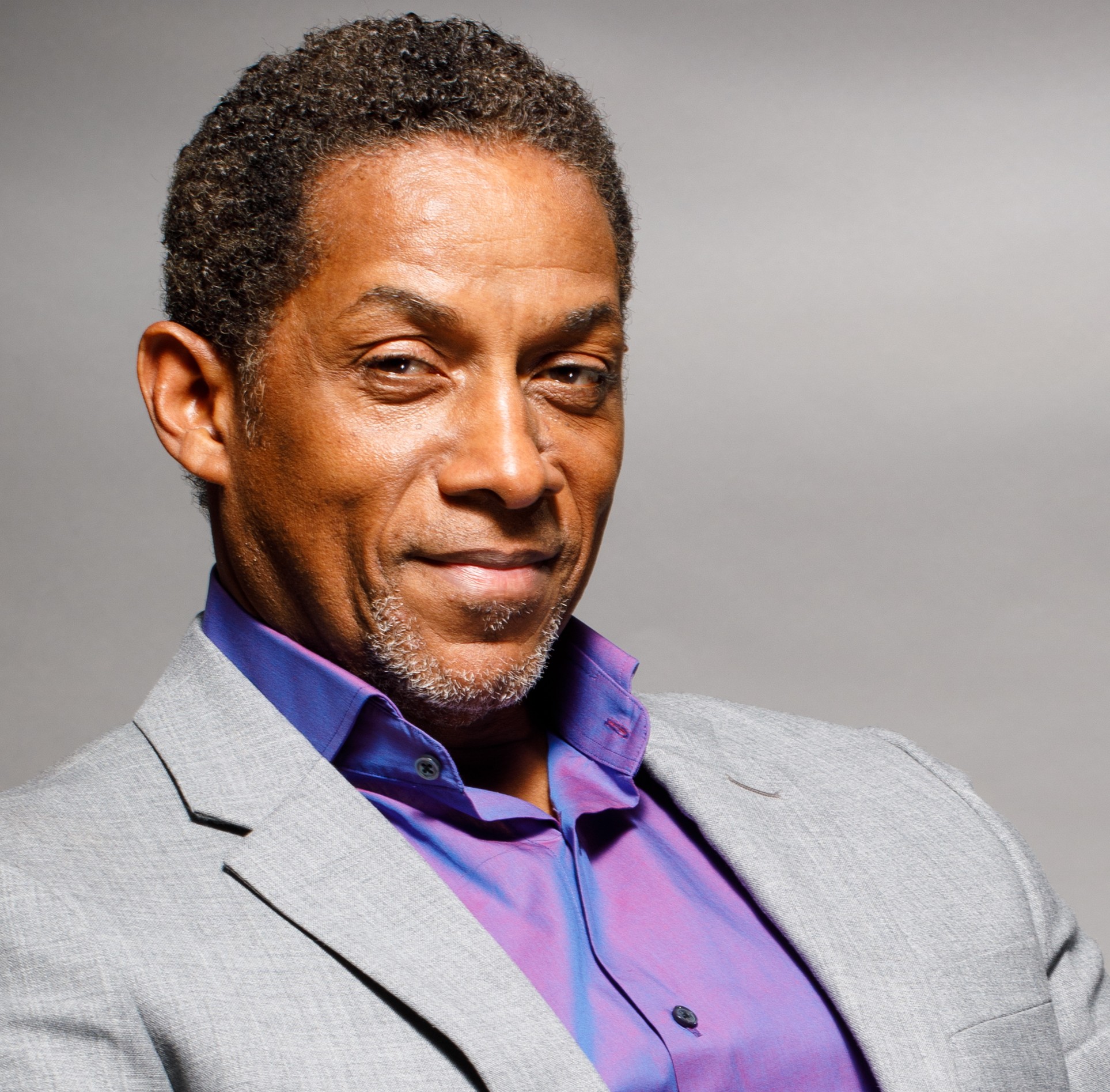 Man with short curly hair and a confident expression, wearing a light gray blazer and purple shirt, against a plain background.
