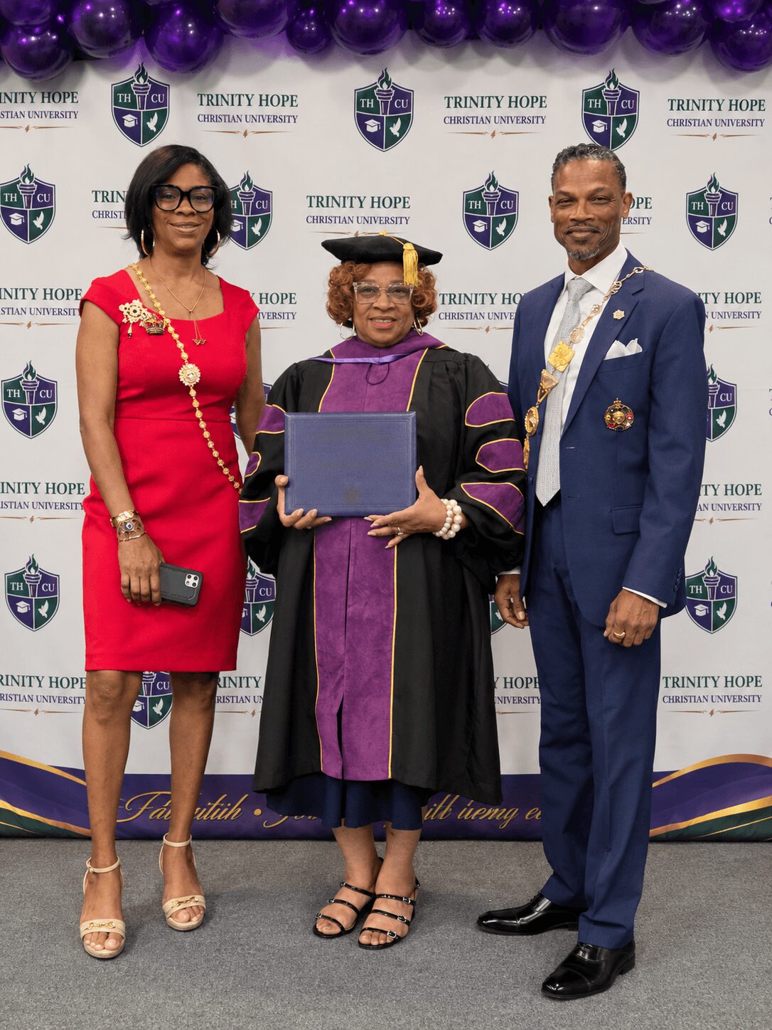 Three people standing in front of a backdrop with Trinity Hope logos, one in graduation attire, two in formal clothing.