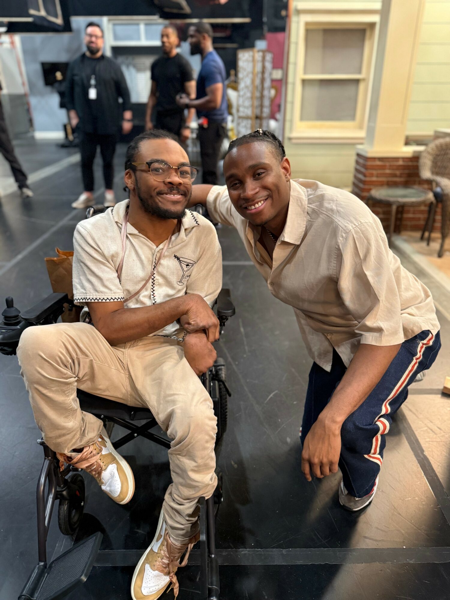 Two people smiling and posing together indoors, one sitting in a wheelchair and the other leaning forward, with a group of people in the background.