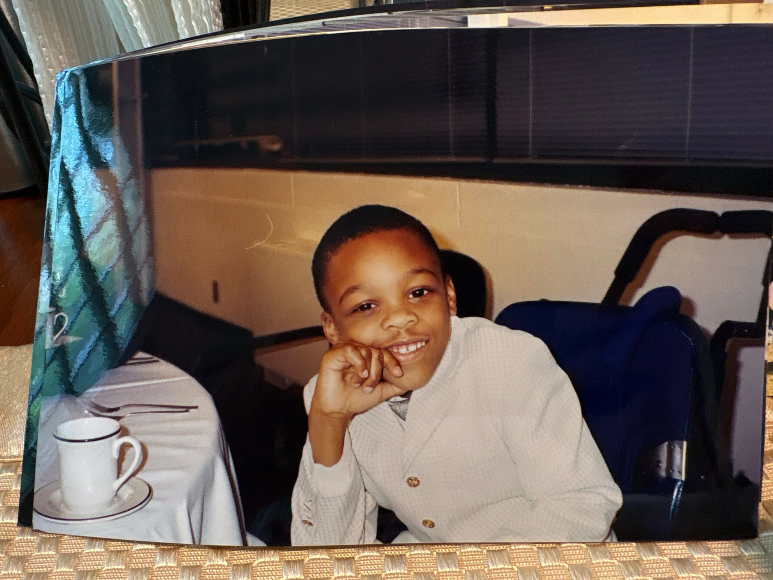 Child smiling with hand on face, sitting at a table with a cup and utensils, in a room with a bed in the background.