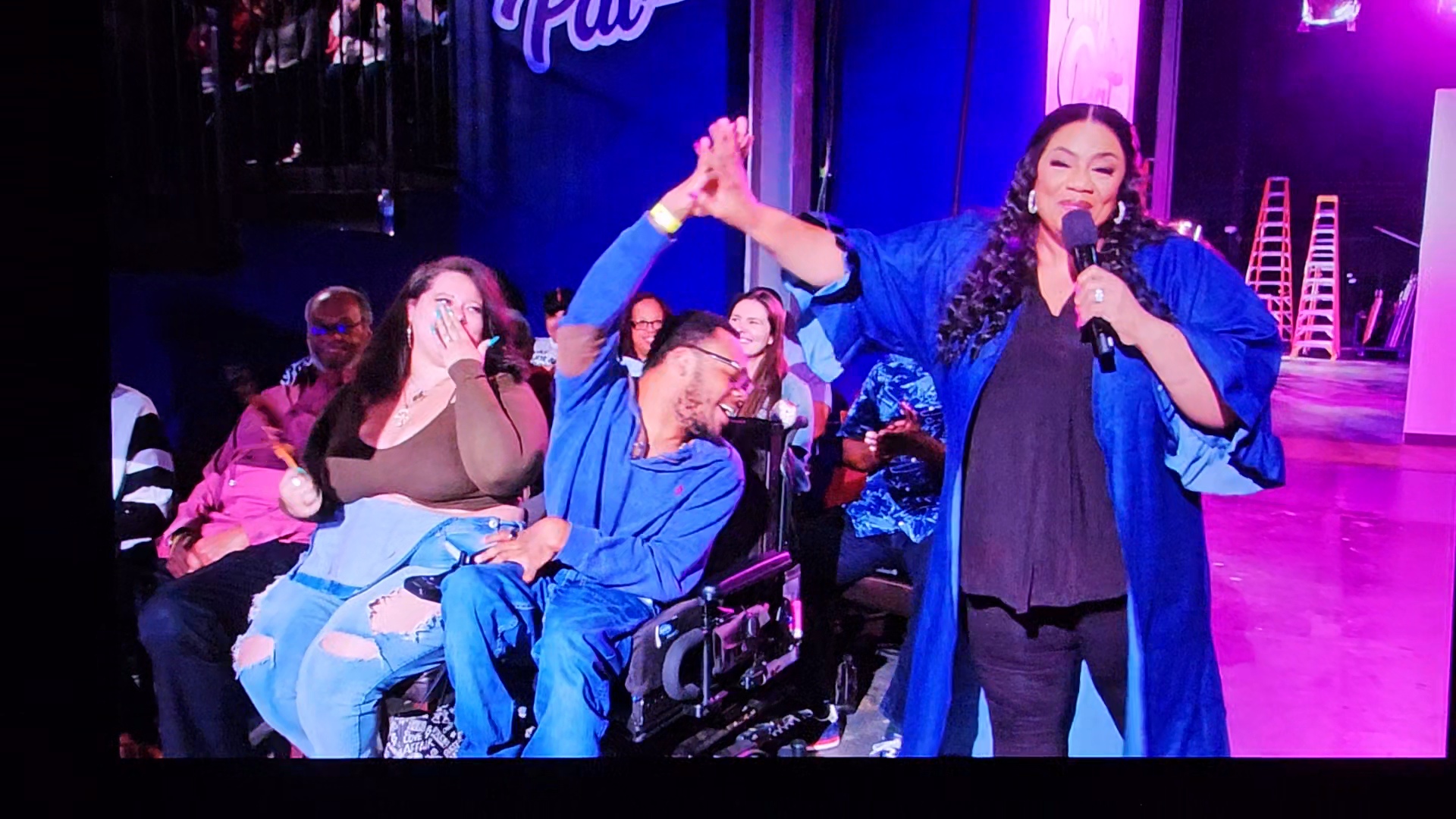 Group of people on stage, one woman speaking into microphone, others clapping and smiling, colorful background, stage lighting.
