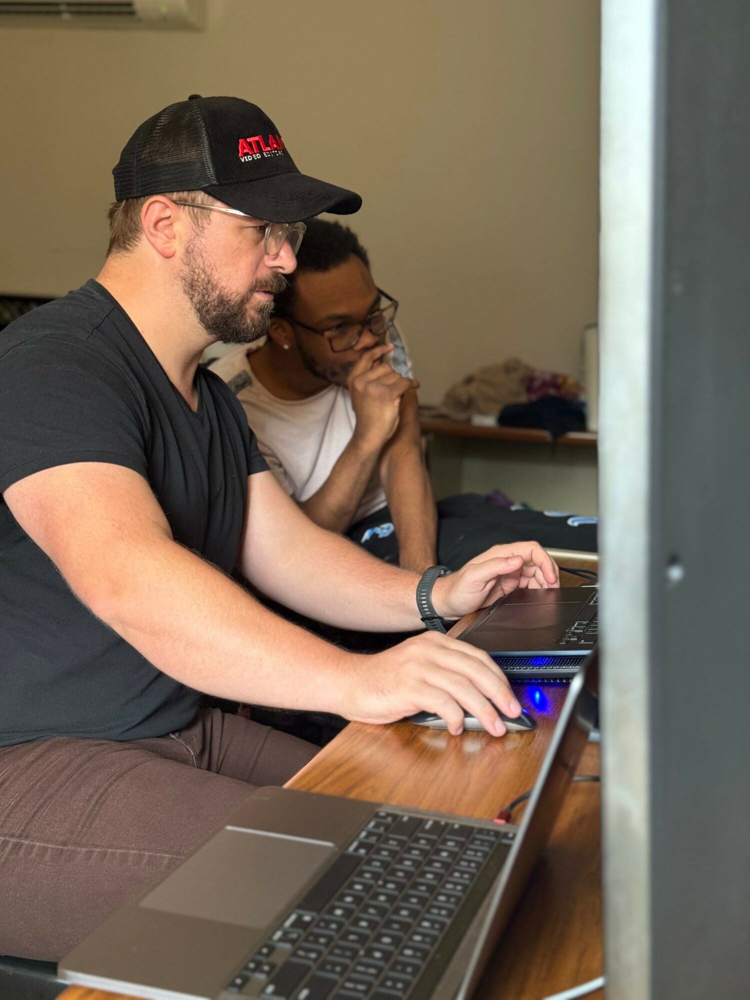 Two people working on laptops at a table, one wearing a black cap and glasses, the other drinking water, in an indoor setting.