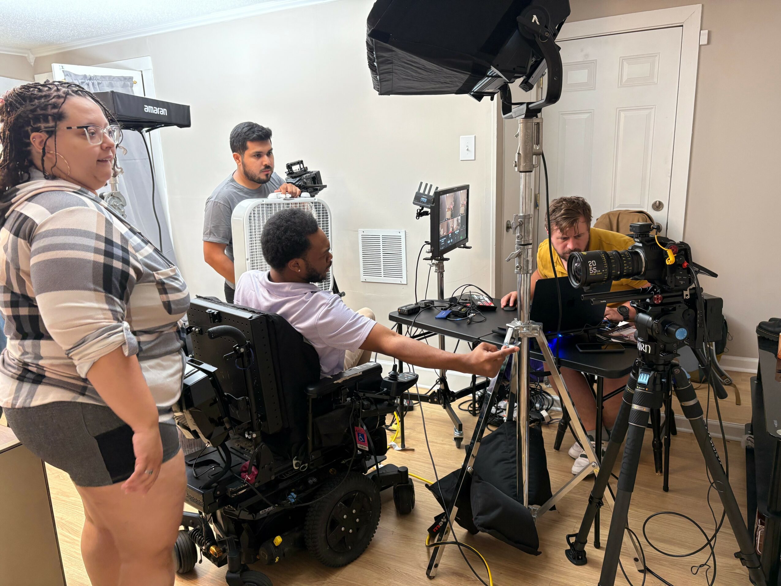 Group of people filming in a room with camera equipment and lighting, some seated and some standing, with a door in the background.