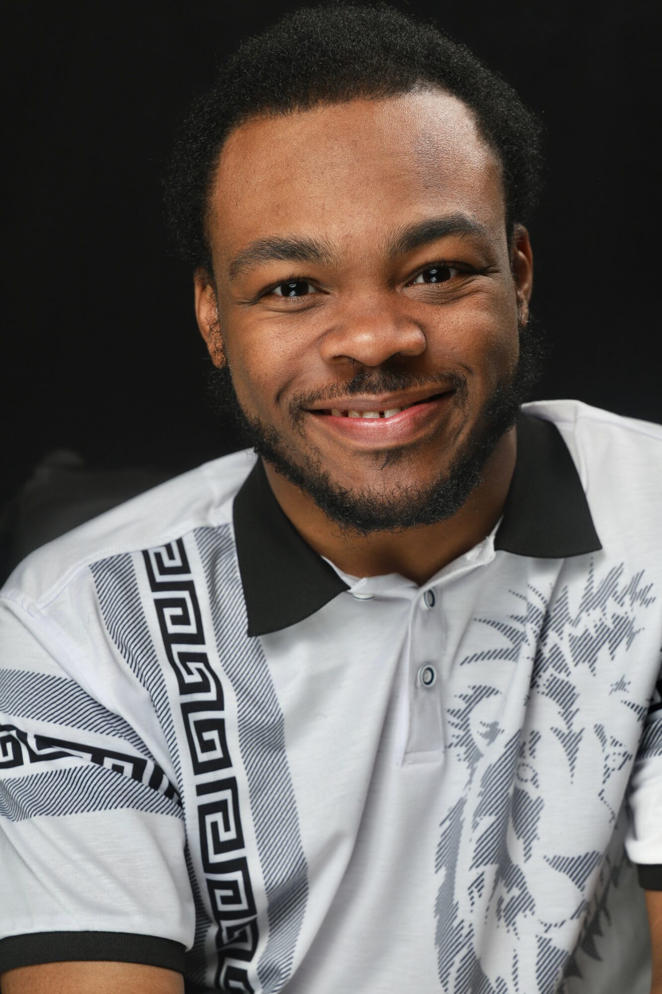 Smiling man with short curly hair and beard, wearing a patterned white and black shirt, against a black background.