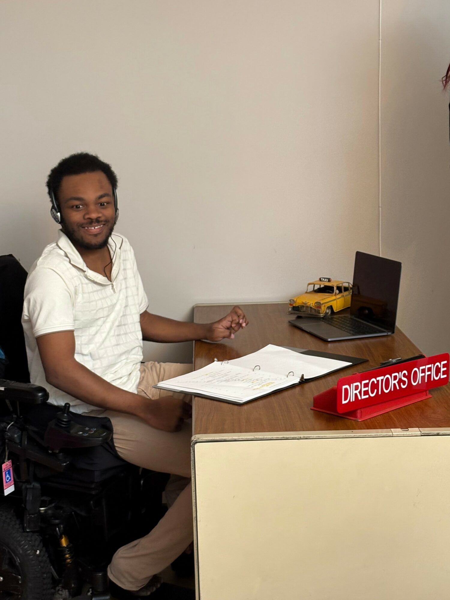 Person sitting at a desk with a laptop, papers, and a red sign reading 'DIRECTORS OFFICE'.