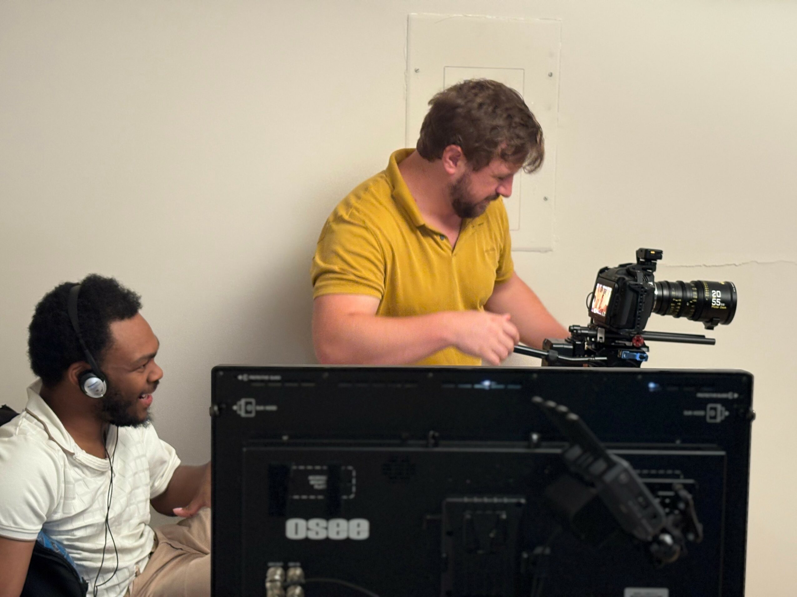 Two men working with camera equipment in a room, one standing and one sitting, both wearing headphones.