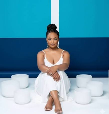 Woman in white dress sitting on a blue bench with white cylindrical stools around her, against a blue background.