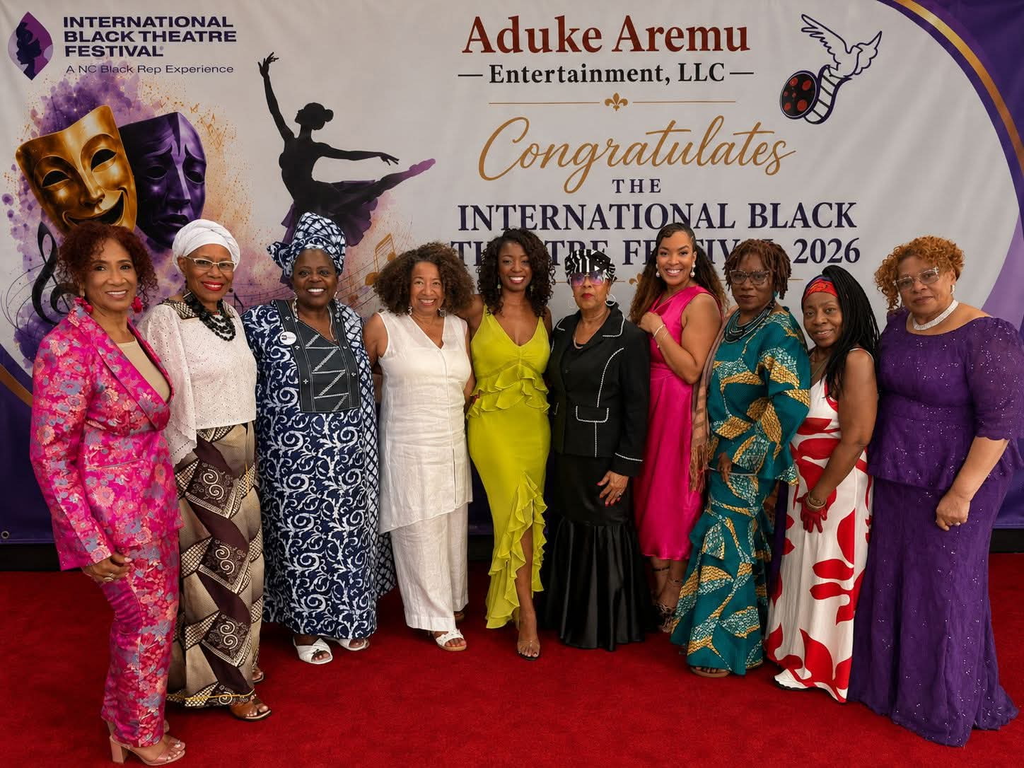 Group of ten women in colorful dresses standing on red carpet at event, smiling, with banner in background. Word 'congratulates' visible.