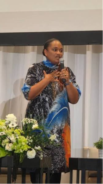 Woman speaking into microphone, standing behind flowers, in front of a white curtain, wearing a patterned dress.