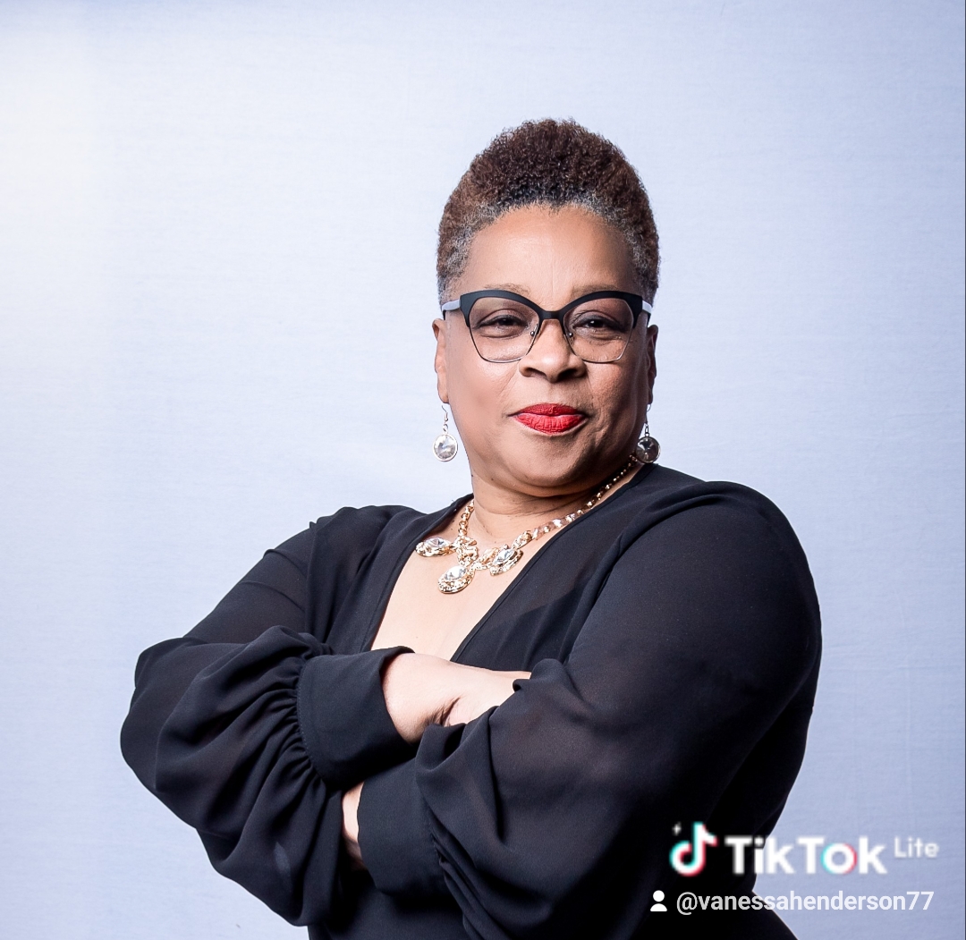 Woman with glasses and jewelry, arms crossed, wearing a black top, standing against a light background.