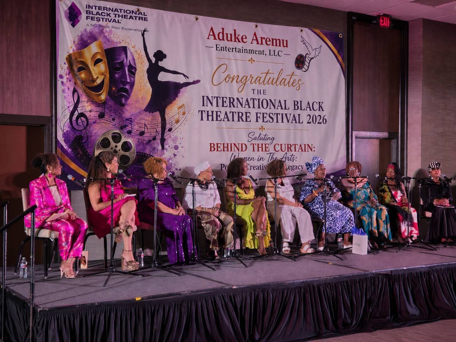 Group of people seated on stage at a theatre festival, with a large banner behind them displaying event details.