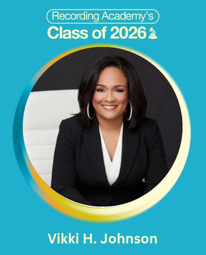 A woman with dark hair and hoop earrings, smiling, wearing a black blazer, seated against a black and white background.