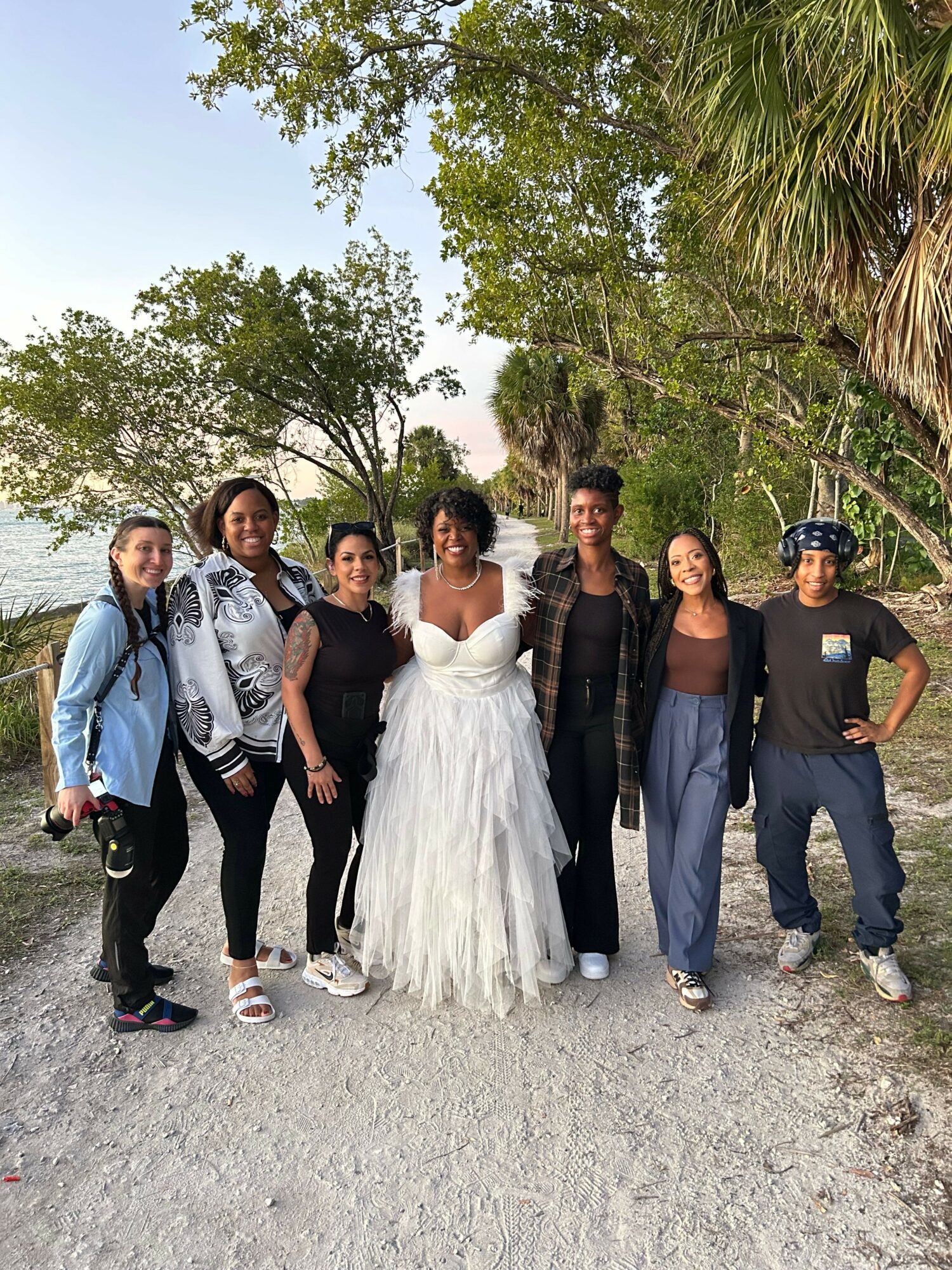 Group of seven people standing outdoors on a gravel path with trees and greenery in the background.