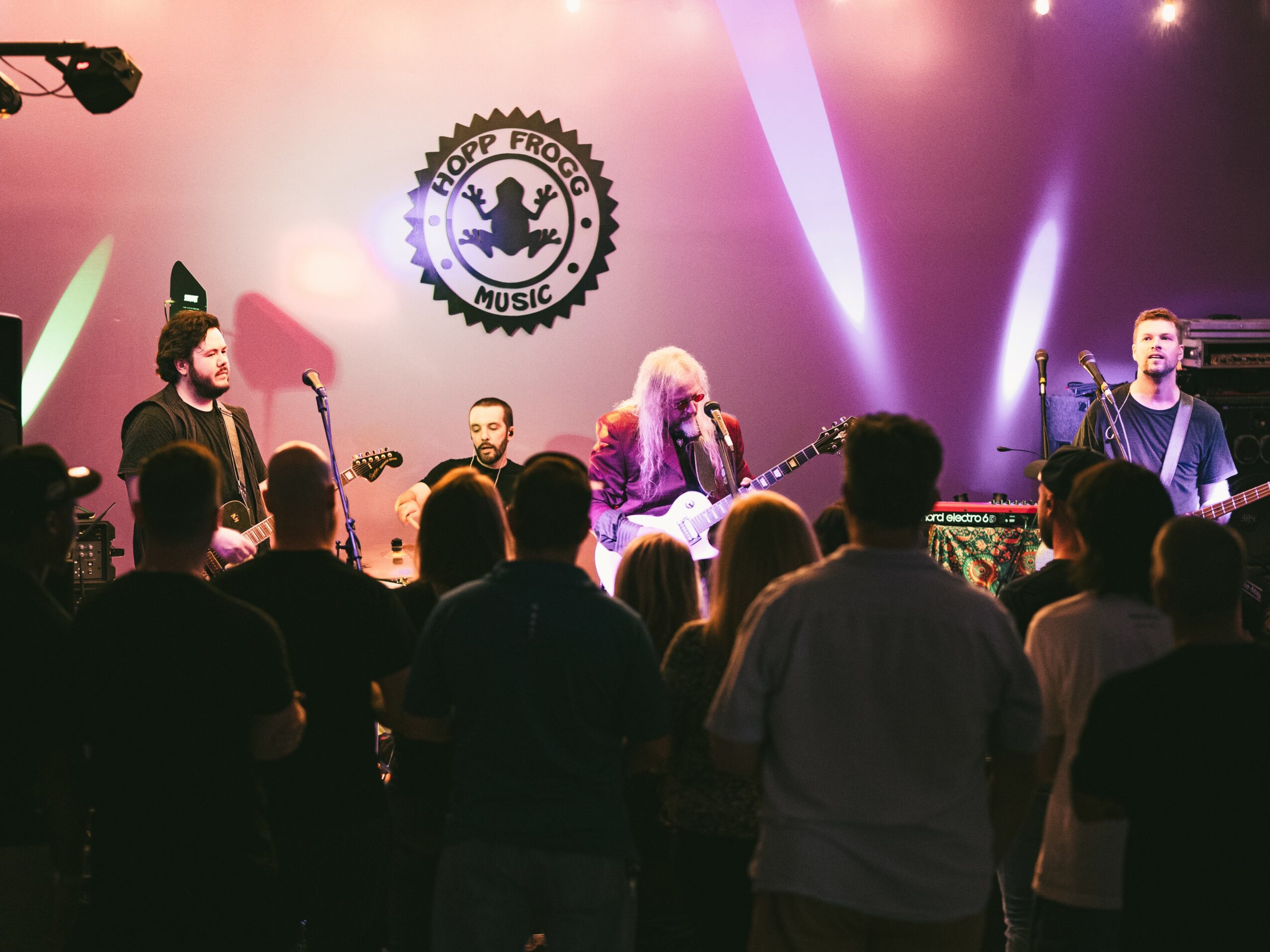 Band performs on stage with audience watching, logo in background, colorful stage lighting, and silhouettes of crowd.