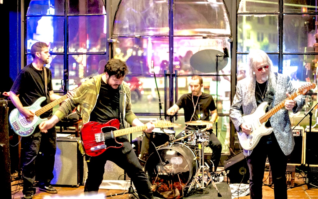Four musicians perform on stage with colorful background lights, playing guitars and drums.