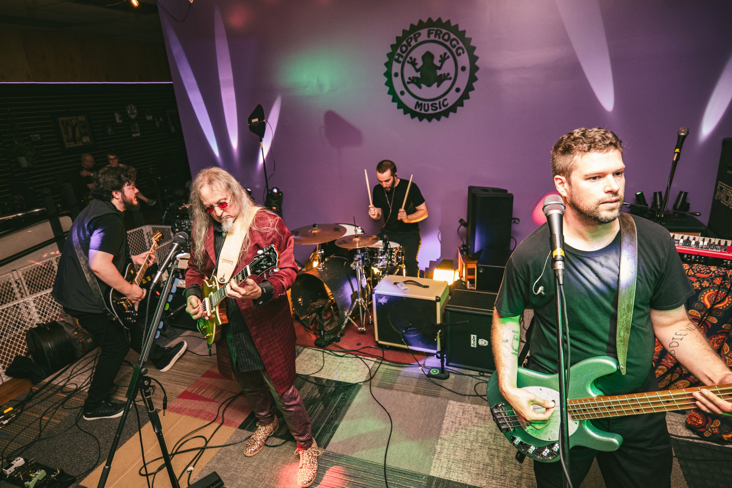 Band performing on stage with purple backdrop and logo, four musicians playing guitars and drums, one singer with blonde hair, stage equipment visible.