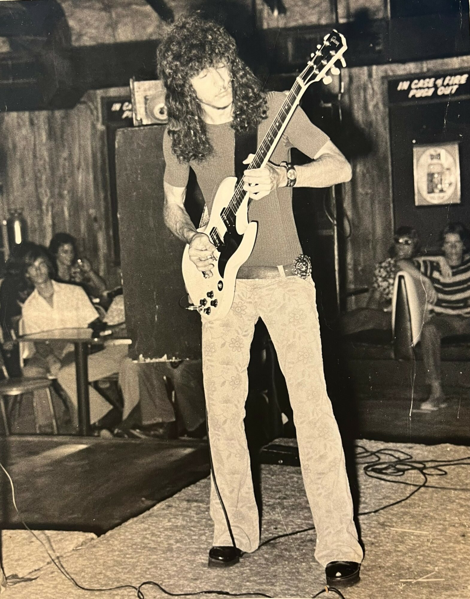 Person with curly hair playing electric guitar on stage, audience seated in background, in a dimly lit venue.