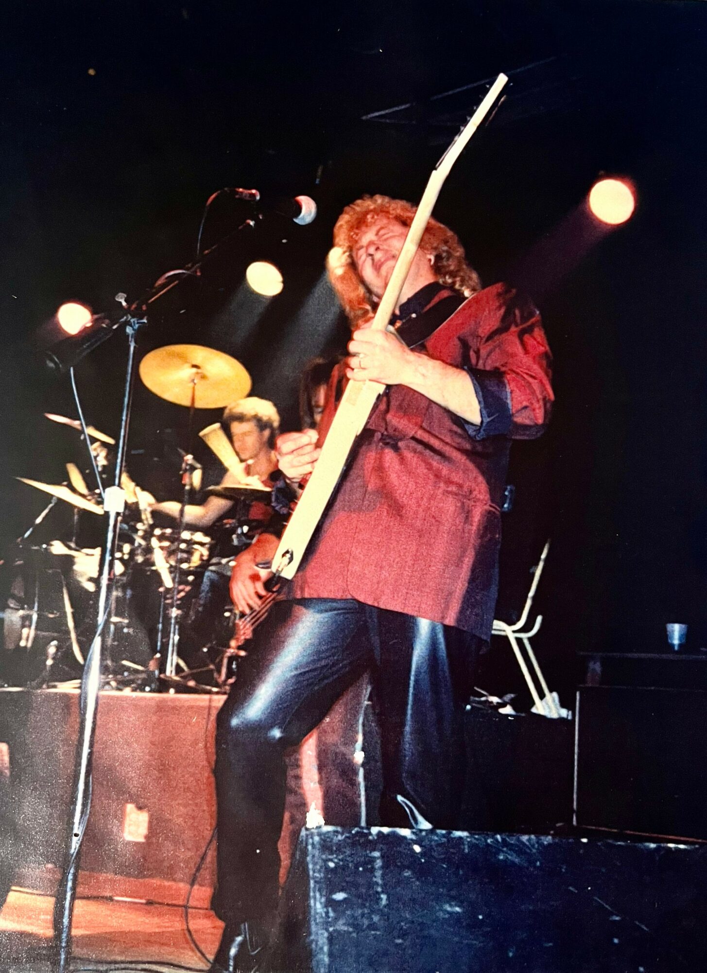 Person playing a large wind instrument on stage with a drummer in the background, stage lights shining down.