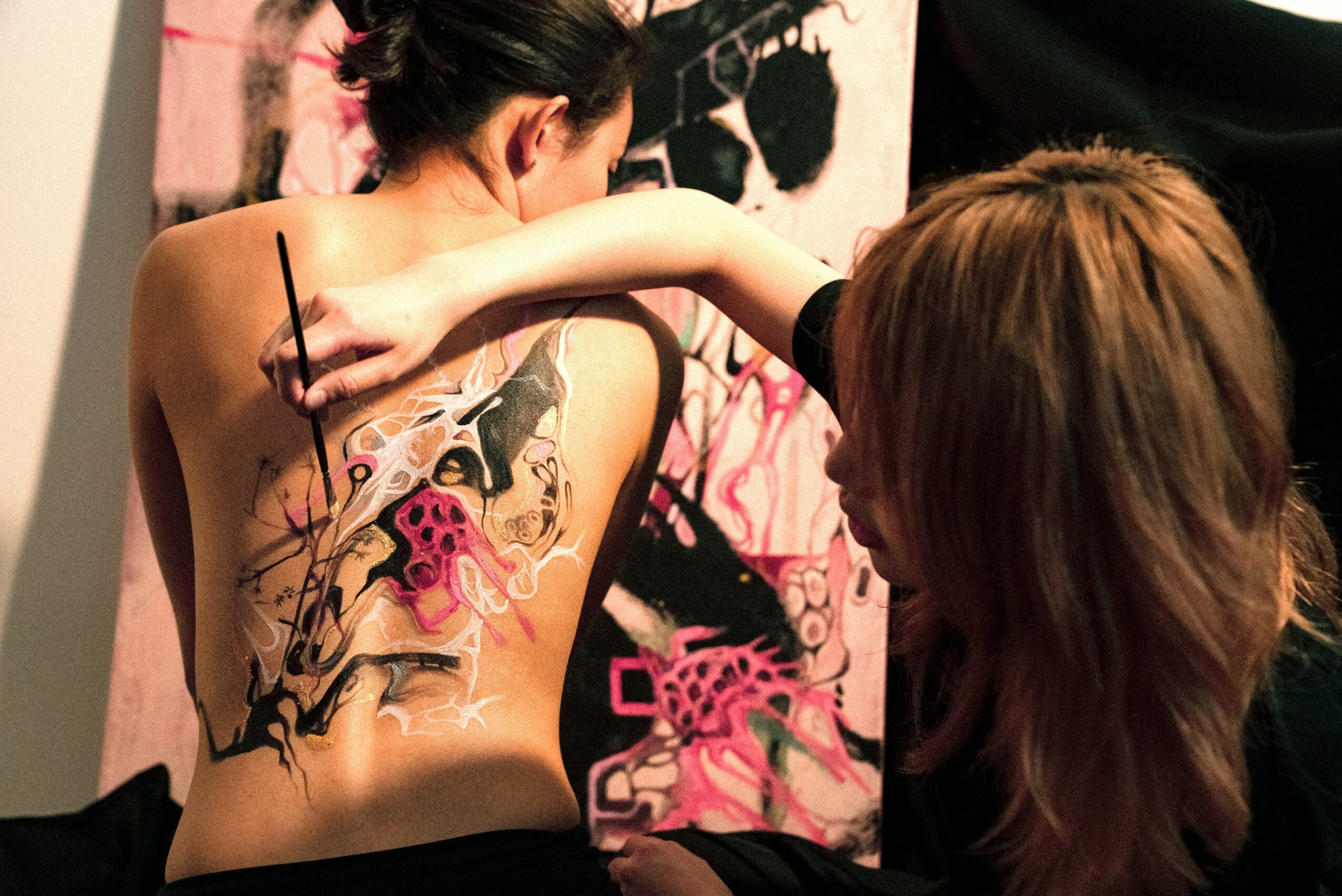 Artist painting a colorful tattoo on a person's back in a studio with pink and black artwork.