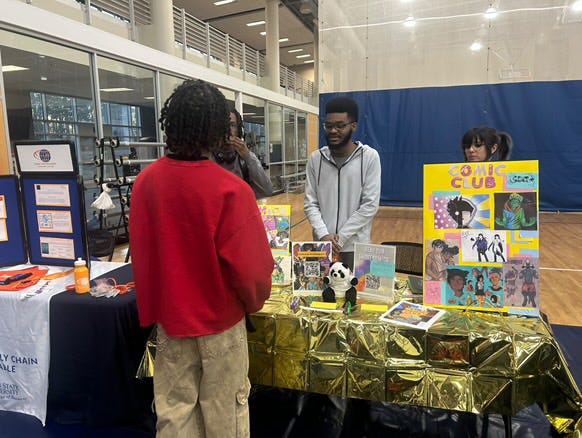 Person in red sweater talking to a person behind a table with colorful displays, in an indoor space.