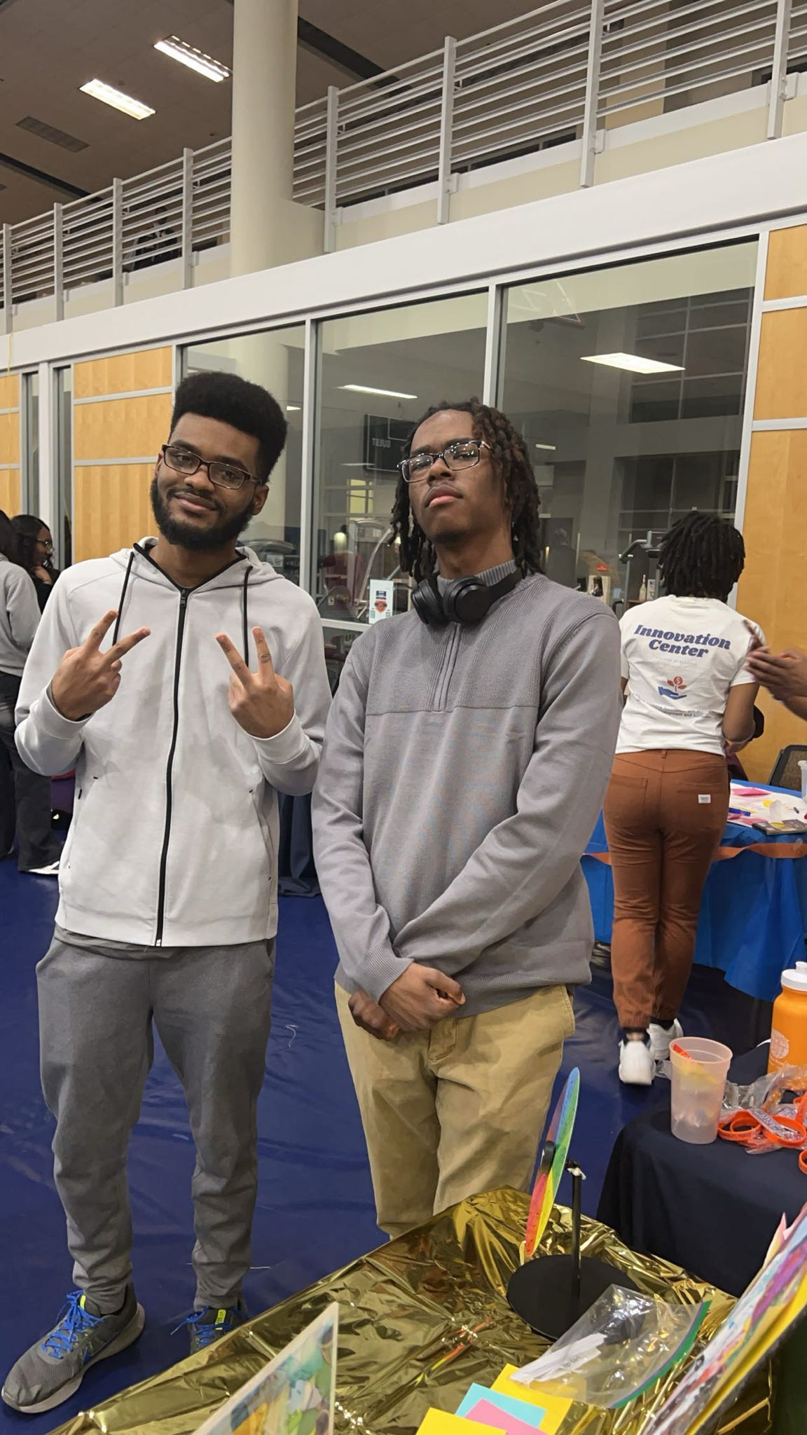 Two young men standing indoors, one smiling and making peace signs, the other with a neutral expression, in front of a table.